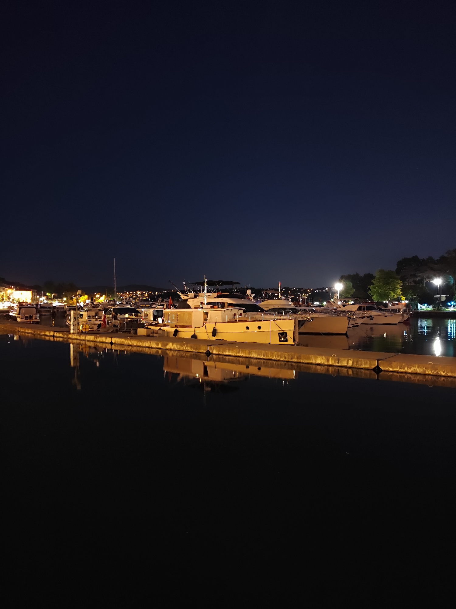 The image depicts a nighttime scene at a marina in the Sarıyer district of Istanbul, Turkey. Several yachts and boats are docked along a concrete pier, illuminated by the warm glow of lights from the boats and nearby buildings. The dark water of the marina reflects the lights, creating a shimmering effect. In the background, the silhouette of trees and hills can be seen against the night sky, with scattered lights indicating the presence of a town or city further inland. A single bright star is visible in the upper portion of the dark blue sky. The overall atmosphere is serene and tranquil.