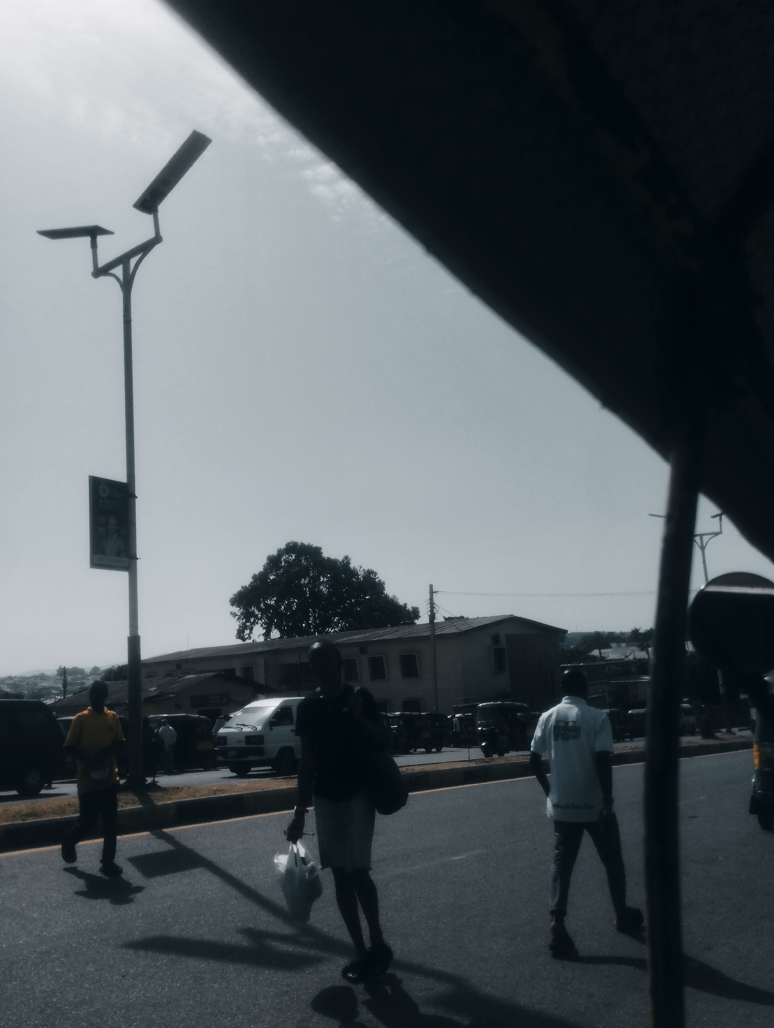 The image captures a street scene in Jos, Nigeria, under a bright, hazy sky. A tall lamppost with a sign attached stands to the left of the frame, its light fixture angled upwards. In the foreground, a paved road is visible with a yellow line marking its edge. Several people are walking on or near the road.
Two men are most prominent in the mid-ground. The man on the left wears a yellow shirt and dark pants, walking with a slight forward lean. He is carrying a dark bag over his right shoulder and appears to be moving away from the camera. The man on the right wears a white short-sleeved shirt with text on the back and dark pants, walking towards the right of the frame. He is carrying a white plastic bag in his right hand.
Behind these individuals, a white van is parked on the side of the road, and various other vehicles, including what appear to be tricycles or small trucks, are visible further down the street. A low-rise building with a tiled roof is situated across the road. A large, dense tree with dark foliage is visible behind the building. The overall impression is of a typical urban environment on a clear day.