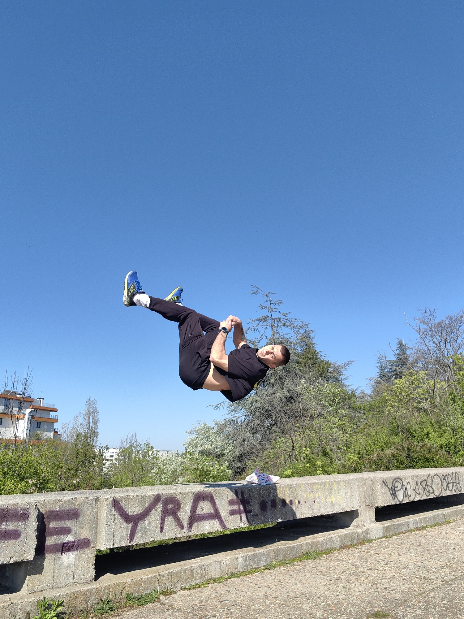 A man is performing a backflip in front of a concrete structure with graffiti in an urban area. The man is wearing a black t-shirt, black pants, white socks, and blue and green running shoes. The sky is clear and blue. In the background are trees and buildings. The graffiti on the concrete structure includes the letters "E," "Y," "R," and "A" in purple paint.