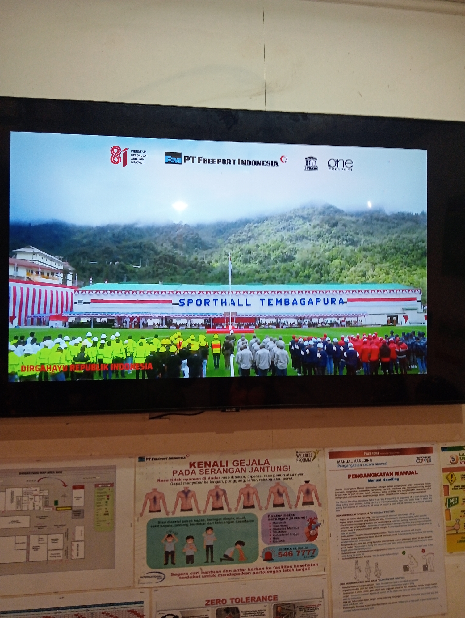 The image shows a large flat-screen television displaying an outdoor event. In the foreground of the televised scene, a large crowd of people are gathered on a grassy field. Many are wearing bright yellow or green vests and white helmets, suggesting they are workers or participants in a work-related event. Behind them, a long building with red and white striped decorations and the words "SPORTHALL TEMBAGAPURA" are visible. The building is set against a backdrop of lush green mountains shrouded in mist. The overall atmosphere appears to be formal, possibly a company event or a ceremonial gathering.
To the right of the television, a wall is visible with several posters displayed. One poster on the far right has a red circle with the number 5 inside and text in a foreign language. Another poster features illustrations of human figures and text related to health symptoms and emergency contact information. A third poster is a map of an area with various buildings and designations. The posters suggest an industrial or operational environment, possibly related to the company "PT FREEPORT INDONESIA" which is also displayed on the television screen. The location is likely within Indonesia, given the language on the posters and the mention of "REPUBLIK INDONESIA" on the television.