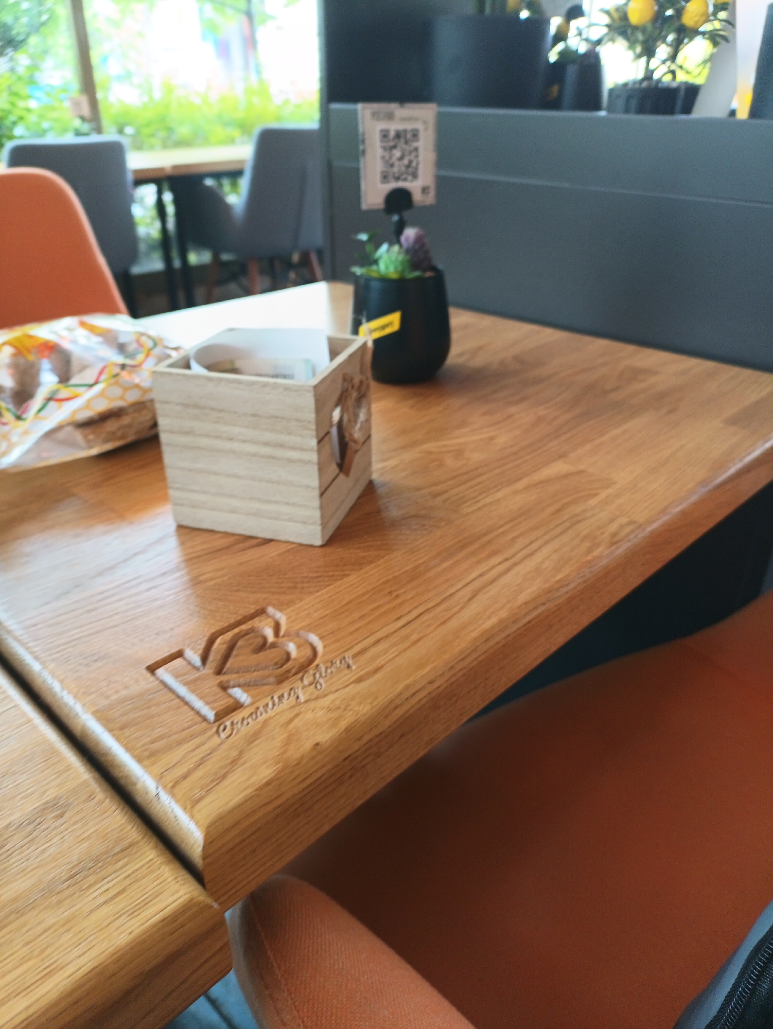 A high-angle shot shows a section of a wooden table with a clear, glossy finish. At the edge of the table is a logo with the letter "K" intertwined with a heart shape, and the words "Creative Glory" inscribed beneath. A square, light-colored wooden box containing folded paper napkins sits on the table. Next to it, there's a partially visible package of food wrapped in clear plastic with a yellow and white patterned design. A small, black plant pot with a single succulent is situated near the edge of the table, accompanied by a QR code stand behind it. The background features a blurred seating area, with gray chairs and hints of a window with greenery outside. The location may be Burgas, Bulgaria but cannot be confirmed from the image.