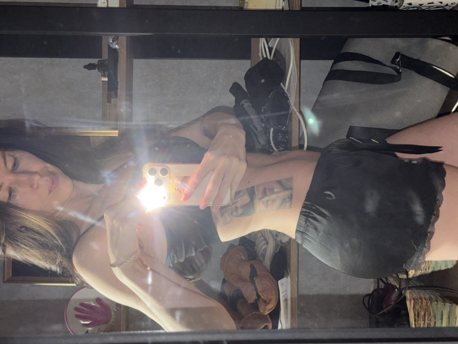 A young woman poses in a mirror selfie, holding her phone, with the flash obscuring her face. She is dressed in black lingerie or shorts with lace trim. Her hair is long and light brown. A tattoo is visible on her side. Behind her, a shelf cluttered with items, including a rolled-up yoga mat, is visible. A small mirror with a hand design is also visible. The reflection includes other objects adding to the cluttered appearance of the space.