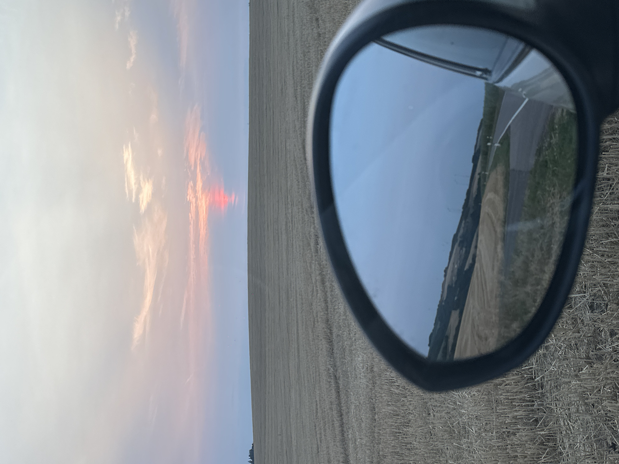 A side-view mirror of a vehicle reflects a landscape under a twilight sky. The mirror shows a paved road curving through rolling hills with fields of stubble and some green vegetation. In the distance, there appears to be a treeline and possibly some structures on the horizon. The sky in the reflection is a deep blue.
The original image shows a view through the side-view mirror, which is framed by the black housing of the mirror. The mirror's reflection captures a scene that is consistent with the exterior view, but it is viewed in reverse. The original image also shows the foreground of a harvested field with stubble. The sky in the original image displays a soft gradient of colors, with hues of pink and orange near the horizon where the sun is setting, transitioning to a pale blue and white overhead. The sun itself is a vibrant pink-red streak just above the horizon. The field in the foreground of the original image is a flat expanse of dry, yellow-brown stubble, suggesting a recently harvested crop, possibly wheat or barley. In the far distance, a dark line of trees or a small cluster of buildings can be seen against the horizon. The overall setting appears to be rural, possibly in the countryside surrounding Ammeldingen bei Neuerburg, Germany, given the visual characteristics of agricultural fields and a rolling landscape often found in such regions. There are no people or animals visible in the image.