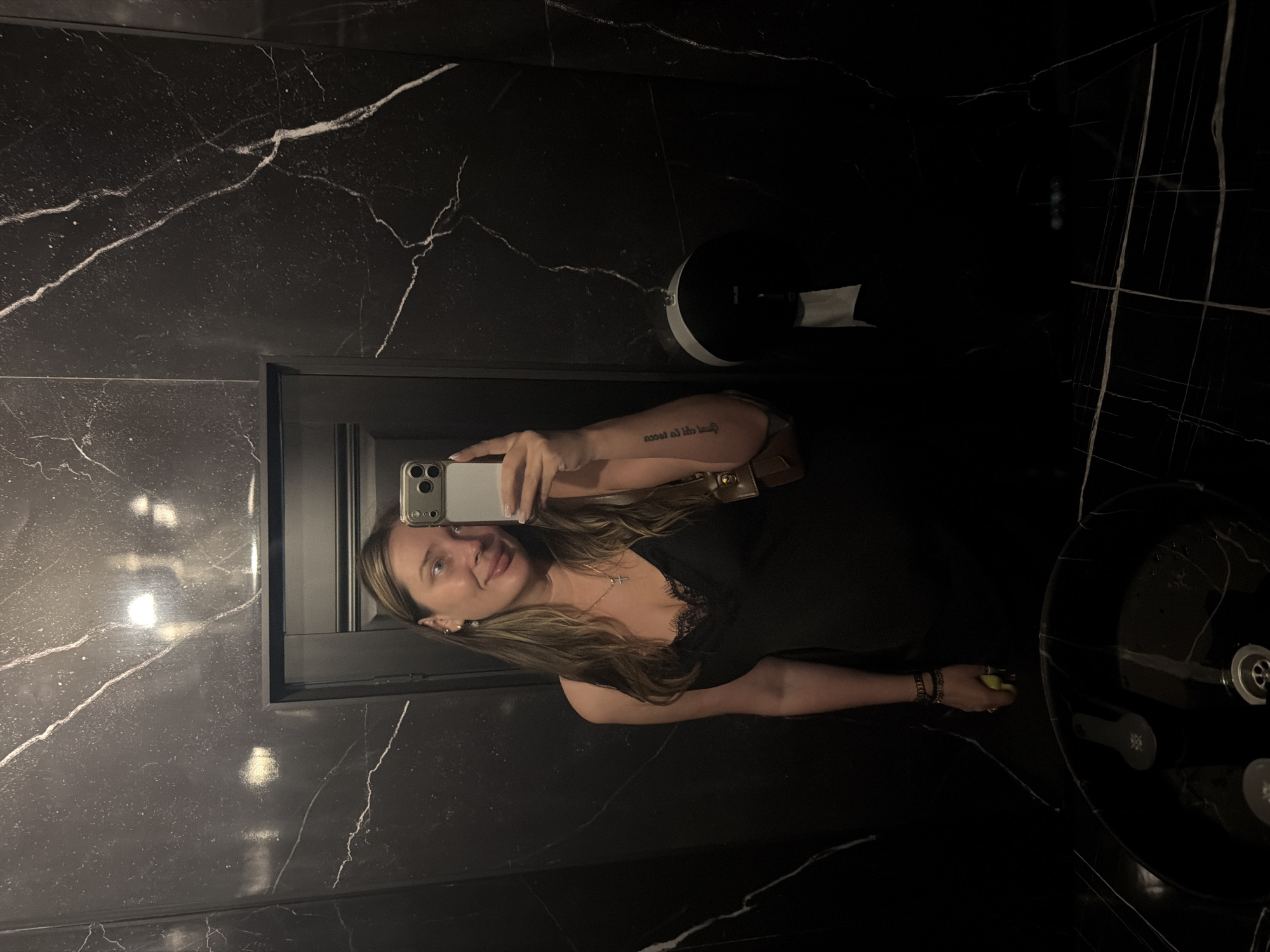 A woman takes a mirror selfie in a dimly lit, modern restroom with black marble walls and a black sink. She is wearing a black slip dress with a lace trim and a cross necklace. A small brown purse is slung over her shoulder, and she has a tattoo on her left arm. Her right hand holds a smartphone to take the picture, and her left hand is holding a yellow object, possibly a banana. The restroom features a soap dispenser and a paper towel dispenser on the wall. The lighting is dim, with reflections from overhead lights visible on the marble surfaces.