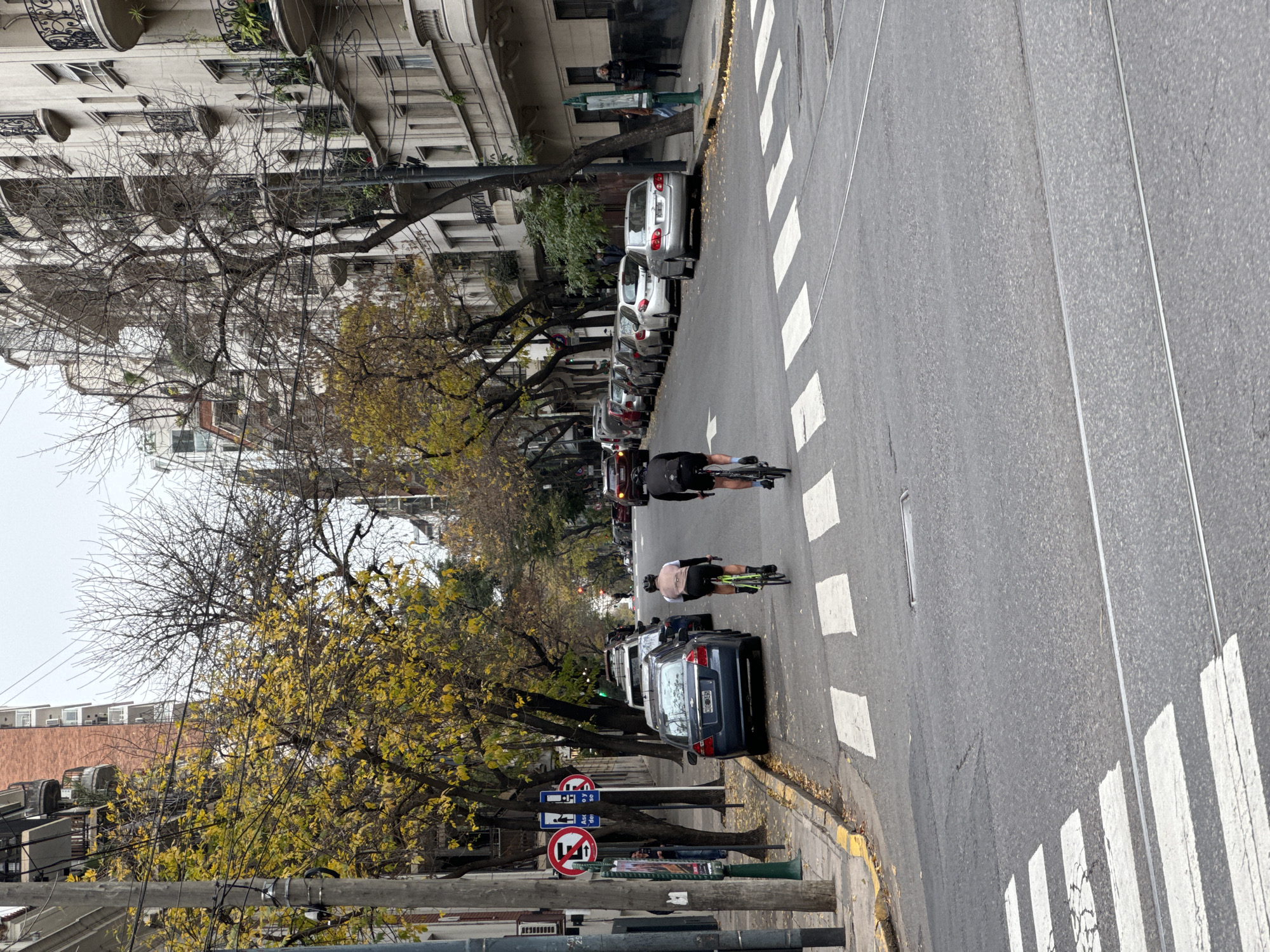 This image depicts a street scene in Buenos Aires, Argentina. The street is lined with parked cars and trees, some of which have yellow leaves, suggesting it may be autumn. Two cyclists are riding down the road. There is a crosswalk in the foreground and background. The buildings on the left side of the street are multi-storied with ornate balconies. There is street signage indicating parking restrictions. The sky is overcast. The presence of streetcar tracks suggests a possibility for the location of the street. Given the architecture and layout of the street, this is most likely Recoleta, Retiro, or Palermo.