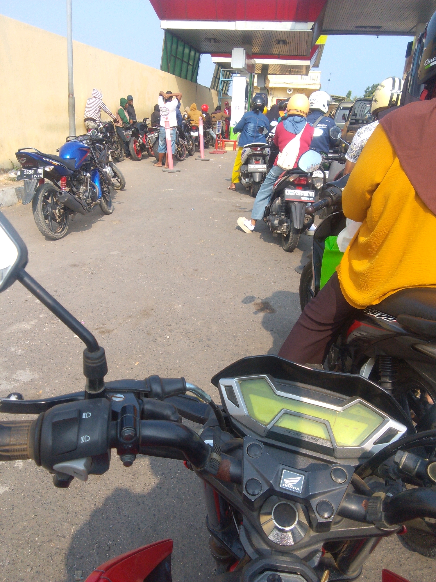The image depicts a scene at a gas station in Semampir, Indonesia, with a clear blue sky overhead and a beige wall lining the left side of the frame. Several motorcycles are parked and in motion, with their riders in various stages of refueling or waiting. The foreground is dominated by the handlebars and dashboard of a motorcycle, indicating the photo was taken from a rider's perspective. The rider in the foreground is wearing a yellow top and dark pants, with a green bag visible on the back of their motorcycle. Other individuals are seen on motorcycles, some wearing helmets. One person in the background is wearing a striped hooded jacket and is near a blue motorcycle. The gas station structure, with a red roof and green accents, is visible in the upper portion of the image. The ground is paved, and a few scattered pieces of debris are present. The overall impression is a busy, everyday scene at a local gas station.