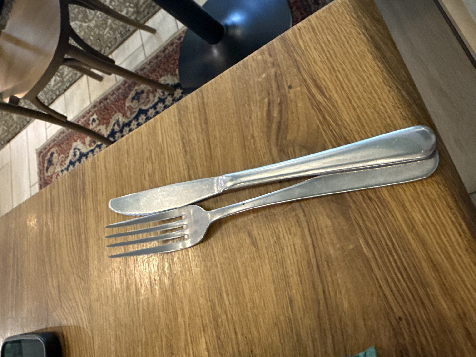 A top-down shot shows a silver fork and knife laid in parallel on a light brown wooden table, likely after a meal. A chair, rug, and table base are visible in the background, adding context to a dining setting. The location is unspecified, but given the prompt it could be Sofia, Bulgaria.