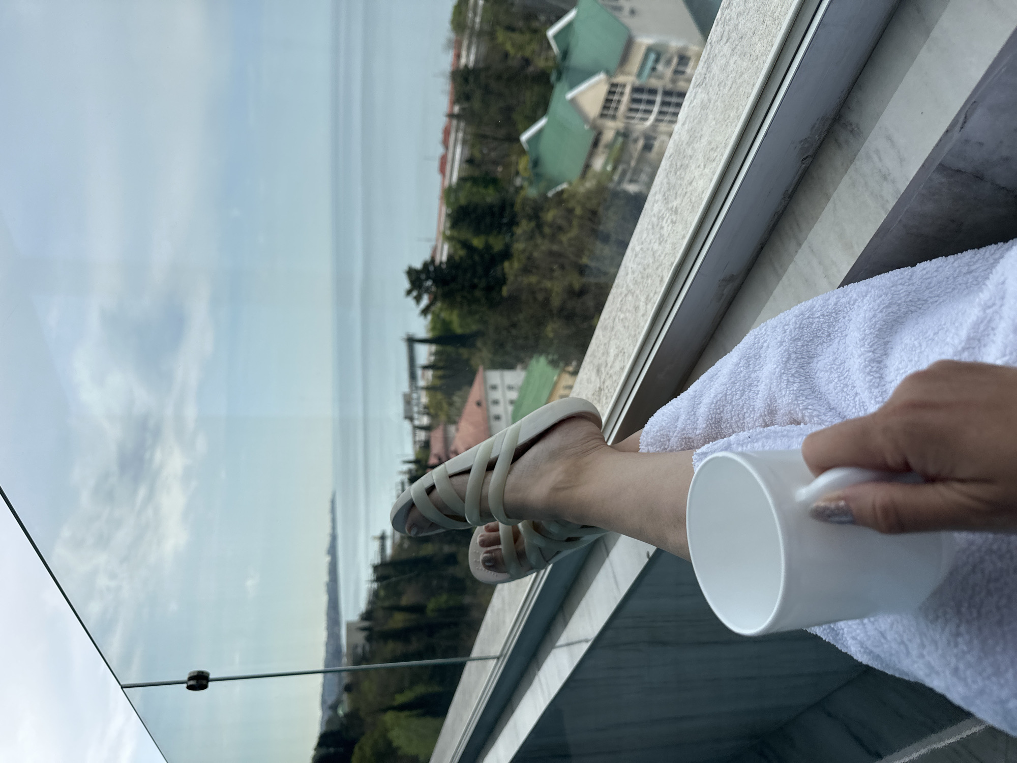 The image presents a first-person perspective from a high vantage point, likely a balcony or a large window, overlooking a scenic view in Sochi, Russia. A person's bare leg, adorned with a light-colored, multi-strapped sandal, is propped horizontally, resting on what appears to be a soft white towel draped over a stone or marble ledge. A hand, with polished nails, holds a simple white mug, suggesting the person is enjoying a beverage.
Beyond the immediate foreground, a transparent glass barrier offers an unobstructed view of a cityscape and natural landscape under a cloudy or overcast sky. In the midground, several buildings with distinctive green roofs are visible, nestled among numerous green trees and lush vegetation. Further in the distance, a large body of water, consistent with the Black Sea in Sochi, stretches towards the horizon, meeting the cloudy sky. The overall atmosphere conveys relaxation and enjoyment of the panoramic view during what appears to be daytime, possibly a late morning or afternoon given the soft light. There is no visible text in the image.