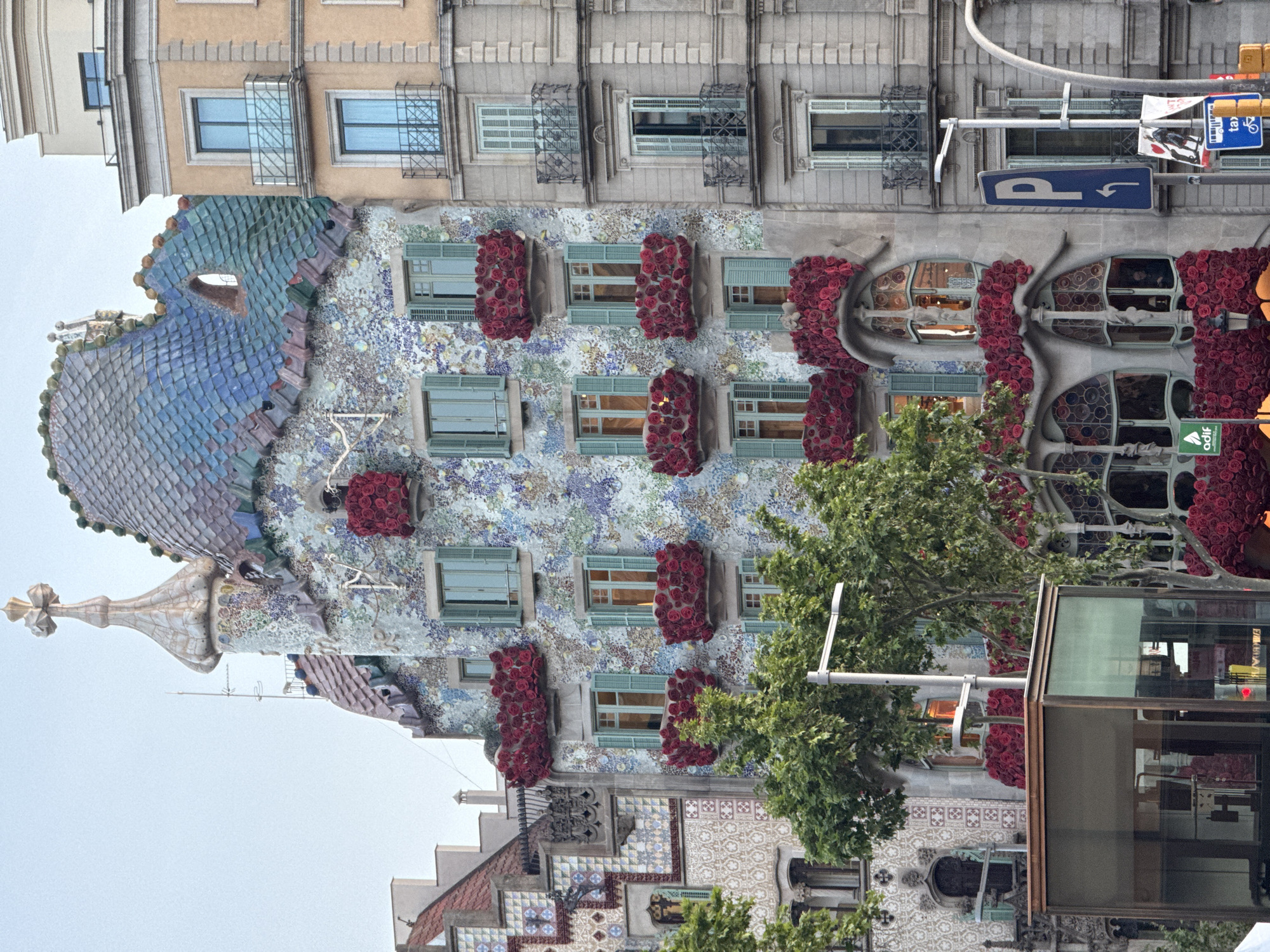 The image captures the vibrant and distinctive architecture of Casa Batlló in Barcelona, Spain, seen from a street-level perspective looking upwards. The building's unique facade is the central subject, characterized by its undulating, organic lines and a mosaic-like surface created from colorful, broken ceramic tiles in shades of blue, green, purple, and beige.
Notable details include the dragon-scaled roof, featuring iridescent blue and green tiles and topped with a prominent, cruciform spire. The building's windows are irregularly shaped, and many feature bone-like supports and balconies adorned with dense, rose-like floral decorations in deep red.
Above Casa Batlló, a more conventional apartment building with rectangular windows and simple balconies with wrought-iron grilles is visible. At street level, parts of a modern glass bus shelter are on the right, and lush green trees partially obscure the lower sections of the building. The bottom part of Casa Batlló transitions to a facade with intricate traditional Spanish or Catalan tile patterns.
The scene takes place during the daytime, under a clear, bright sky. There is no specific activity taking place, rather it is a static urban scene showcasing a famous architectural landmark.
Visible text includes multiple street signs: a blue square sign with a white 'P' and an arrow indicating parking, a "TAXI" sign, and a blue sign with a bicycle symbol. A partially visible green sign with the text "odif" is also present near the street level.
