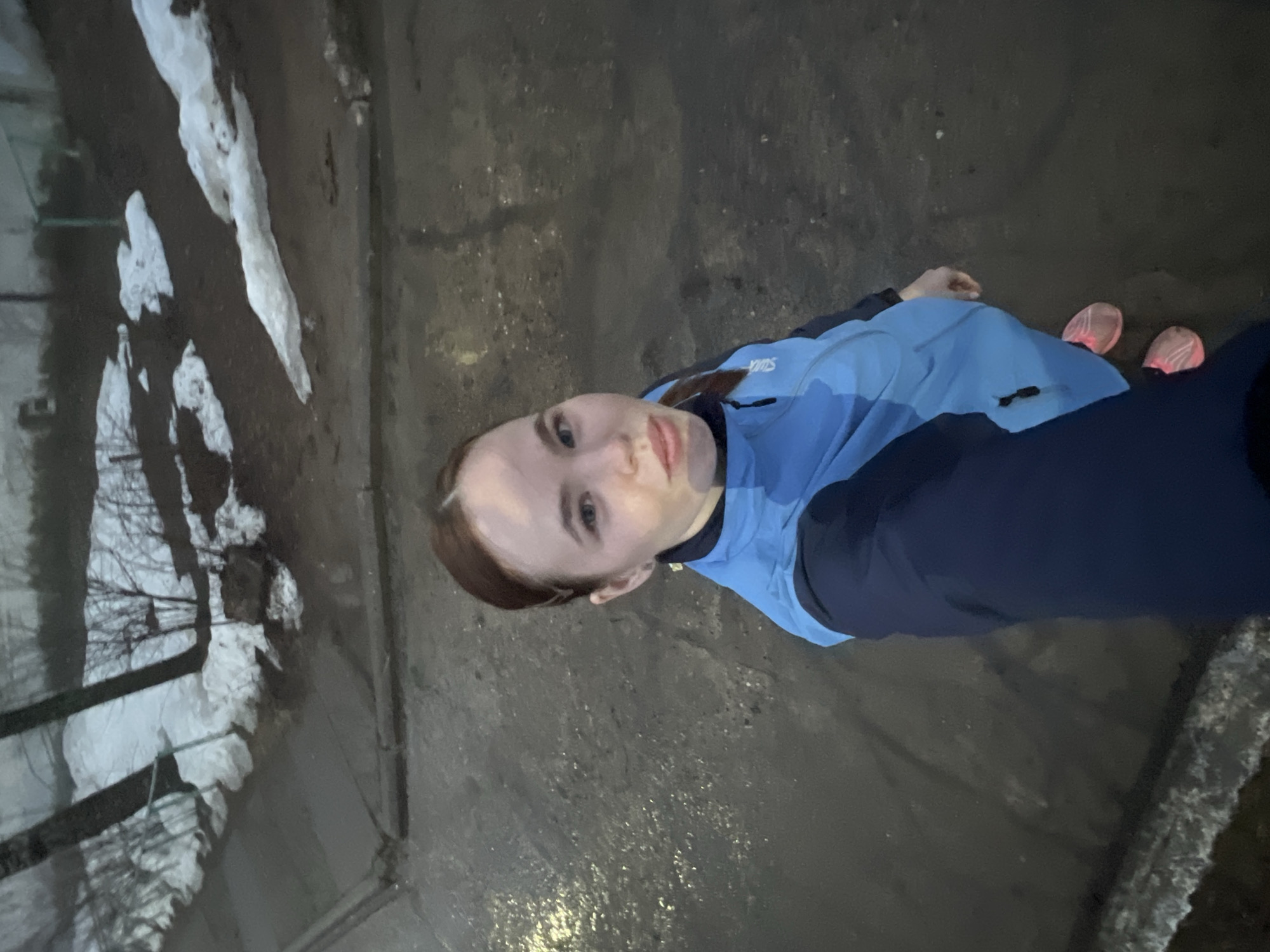 A woman is standing outdoors on a paved surface that appears wet and dark. To the left, there is a curb partially covered in snow and some bare branches visible against what may be a building. The woman wears a blue jacket with dark sleeves and pink running shoes. The overall impression is that she is taking a selfie during or after a run or walk in Chaikovsky, Russia, in cold weather. The image does not provide enough information to confirm the specific location or time of day.