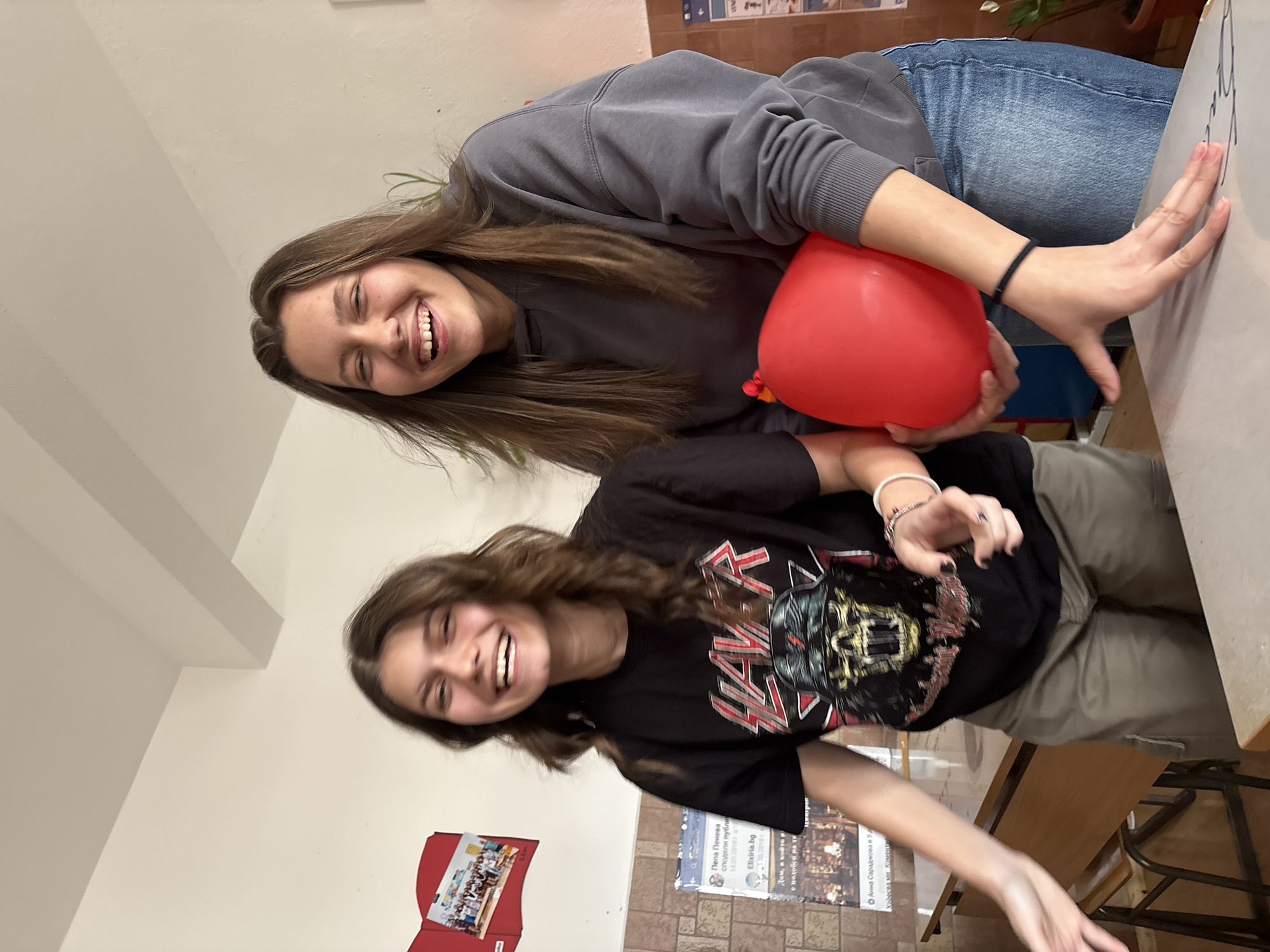 Two young women are smiling for a photo indoors. The woman on the left is wearing a gray sweatshirt and jeans and is holding a red balloon. The woman on the right is wearing a black t-shirt with a band logo and khaki pants. They are standing near a table. There is a bulletin board on the wall behind them with various papers and posters. One of the papers has Bulgarian text on it and mentions "Elixiria.bg". Based on the text on the paper, the location is likely in Bulgaria, possibly Panagyurishte.