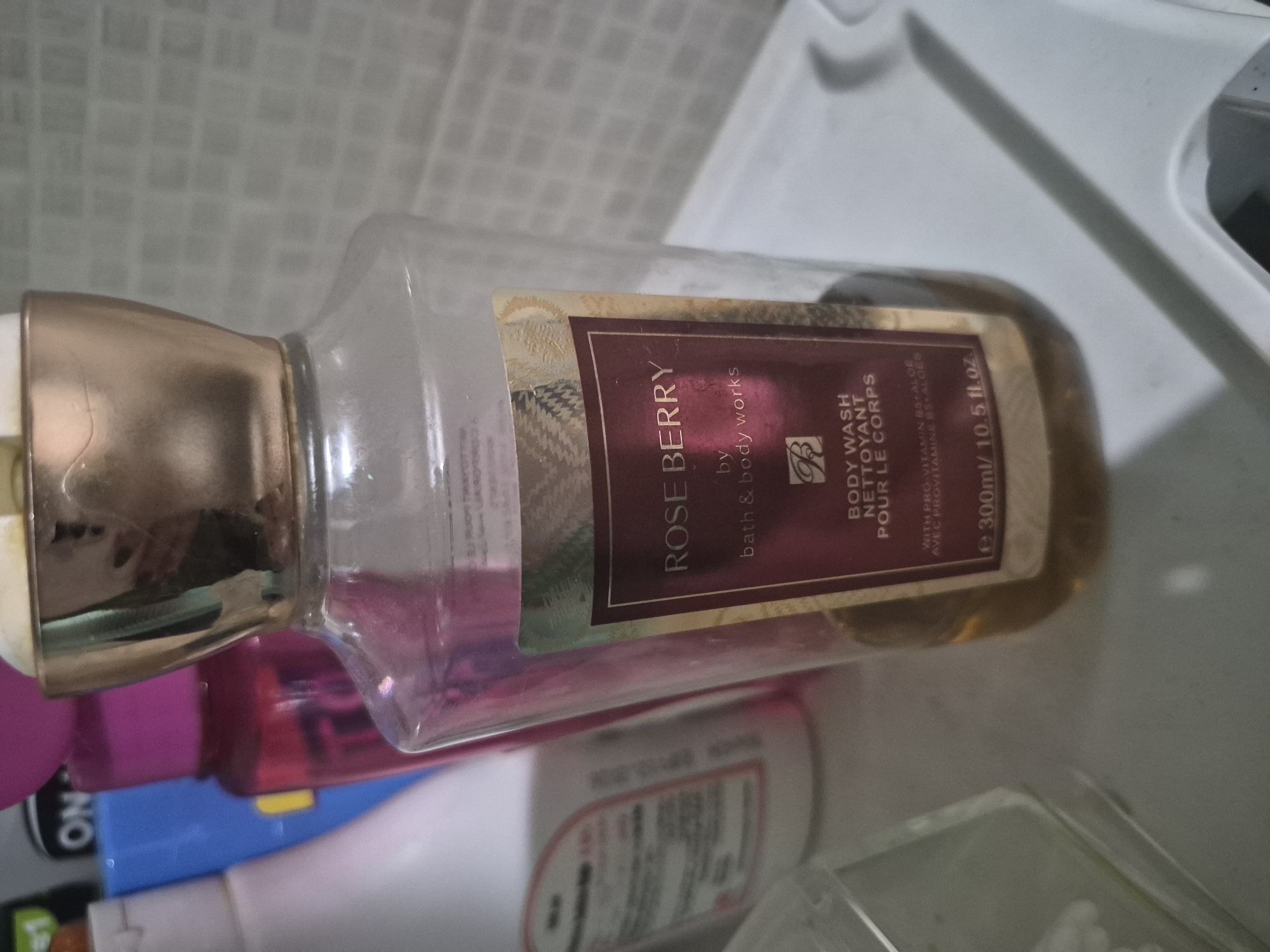 The image displays a clear plastic bottle of body wash, filled with an amber-colored liquid, lying on its side. The bottle has a metallic gold-colored pump or cap. Its label is predominantly dark red or maroon with gold design elements and text.
The primary subject is a Bath & Body Works product, as indicated by the text "by bath & body works" on the label. The product is identified as "ROSE BERRY" and a "BODY WASH / NETTOYANT POUR LE CORPS." Further details on the label state "WITH PRO-VITAMIN B5 ALOE / AVEC PROVITAMINE B5 ALOES." The volume is marked as "e 300ml / 10.5 fl.oz."
The bottle is positioned against what appears to be a light-colored, possibly tiled or textured surface, suggesting an indoor setting like a bathroom or a storage shelf. Partially visible in the background are other personal care products or bottles, including one with "ON" visible on its label, and another with a bright pink color. No people are visible, and no specific activity is taking place; the image is a still life of stored items. The overall context suggests this item is located in Yerevan, Armenia.