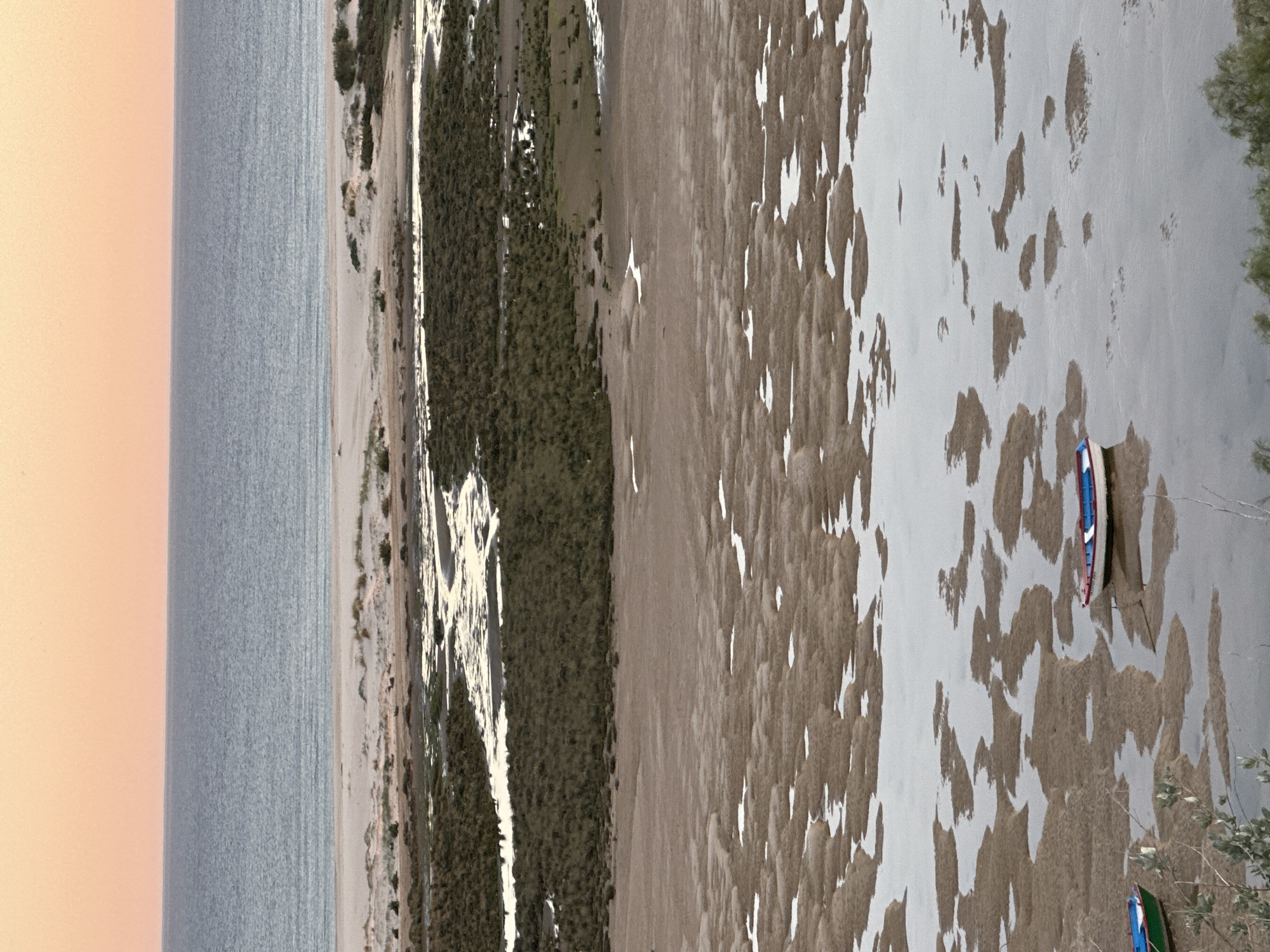 The image depicts a coastal landscape in Vila Real de Santo António, Portugal. The scene includes the Atlantic Ocean on the left with a hazy, orange-tinted sky above. A shoreline composed of sand dunes and sparse vegetation runs parallel to the ocean. The right portion of the image features a large expanse of mudflats or wetlands, likely part of the Ria Formosa Natural Park. These flats are covered with a pattern of brown sediment interspersed with shallow pools of water. Two small boats, one red and one green, are visible near the edge of the wetlands. The overall impression is of a tidal ecosystem at low tide.