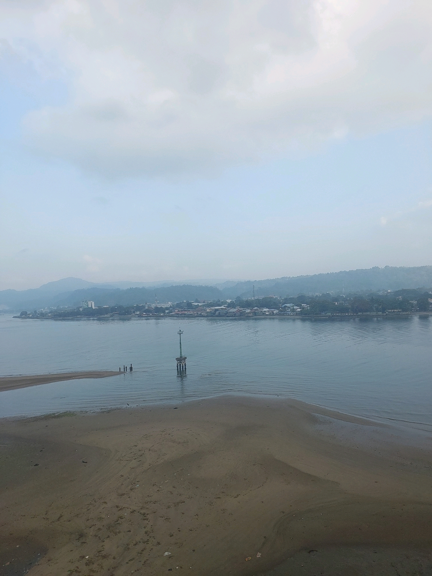 The image depicts a wide, calm body of water with a distant, hazy coastline of Ambon, Indonesia. In the foreground, a sandy, low-tide area is visible with rippled sand formations. A small group of three individuals stands in the shallow water near a sandbar, appearing as dark silhouettes. Further out in the water, a navigational marker or buoy stands on stilts. The background shows a densely populated shoreline with buildings of various sizes, including a prominent, taller building with a green roof, characteristic of urban development. Rolling hills, also shrouded in haze, form the backdrop to the city. The sky is overcast with soft, diffused light, suggesting it might be early morning or late afternoon. The overall scene conveys a sense of tranquility and a typical coastal view of a developed area in Ambon.