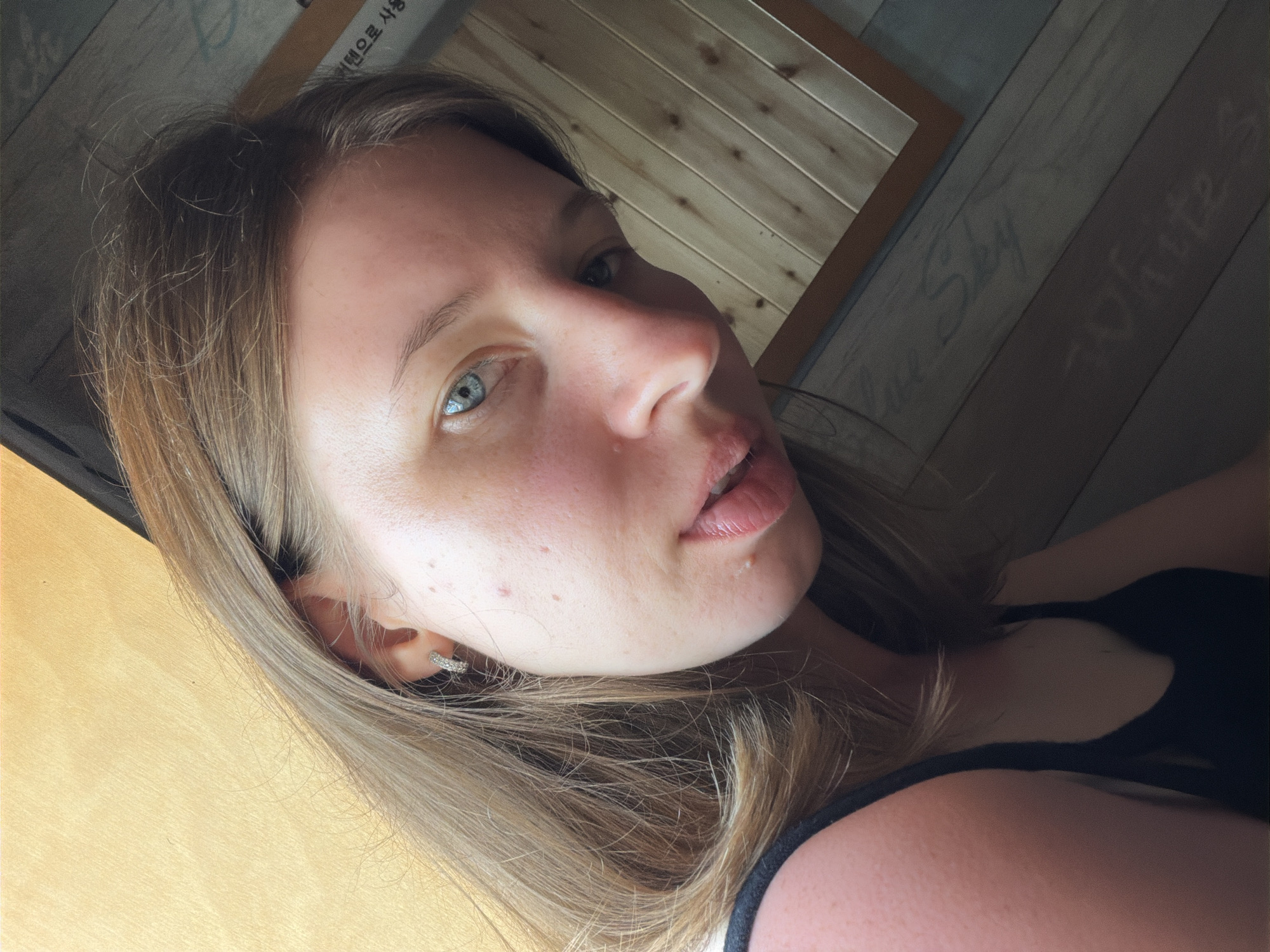 A young woman with light brown hair and blue eyes is pictured in a close-up, slightly angled selfie. She is wearing a black tank top and a small hoop earring in her left ear. Her skin has visible blemishes and freckles. The background consists of wooden walls with a light-colored, textured surface on the left and planks of wood on the right, some of which have faint lettering. This setting appears to be indoors, possibly a cafe or restaurant in Gangneung-si, South Korea, as suggested by the presence of Korean text on the wooden paneling. The lighting is soft and natural.