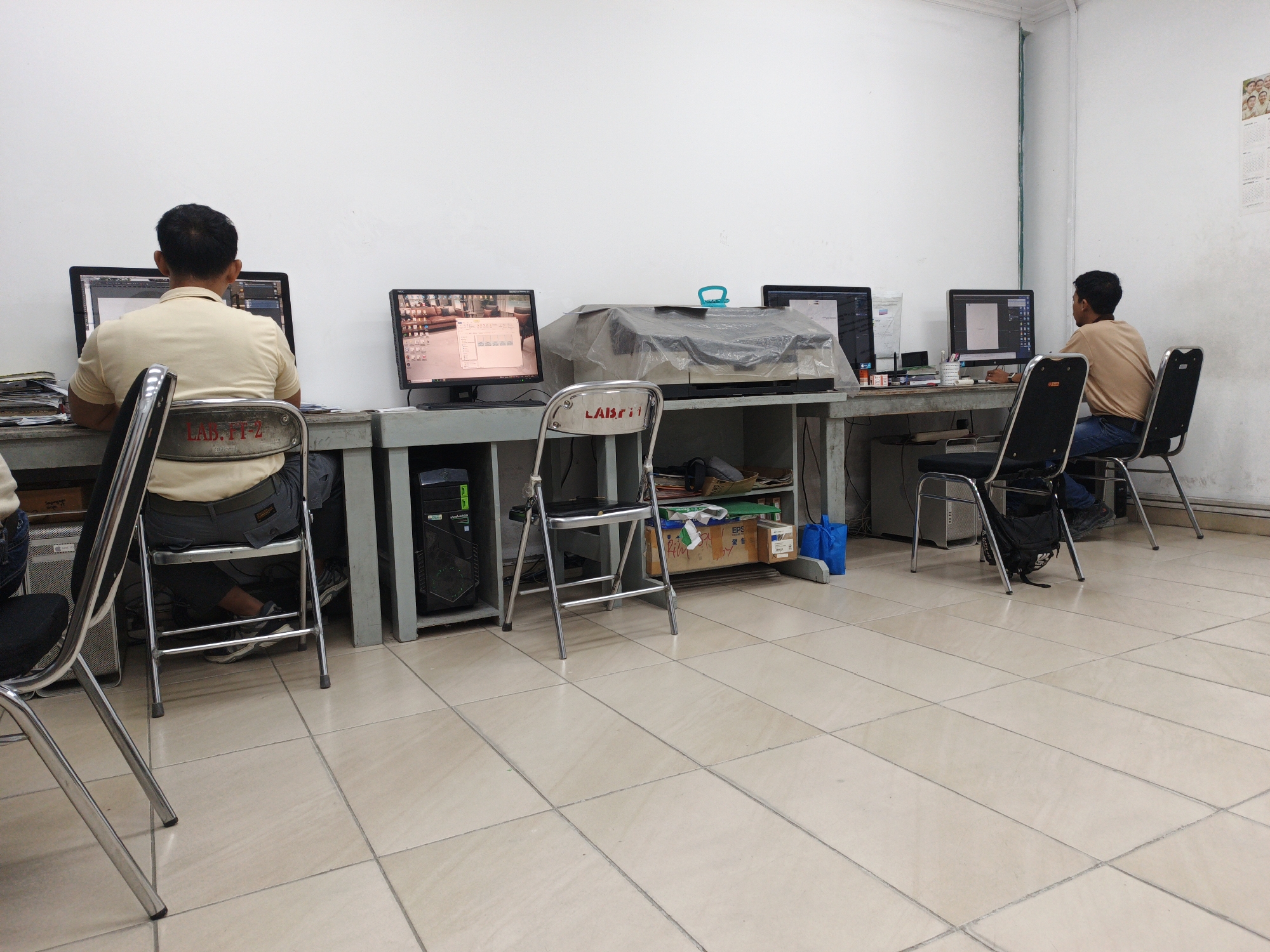 This image shows an interior scene, likely a computer lab or office in Ngoro, Indonesia. Three men are seated at desks, each in front of a computer monitor. The desks are arranged in a row, with computers, keyboards, and other office supplies visible.

In the foreground, the legs of two empty chairs are visible. The man on the far left is wearing a light-colored polo shirt and dark pants, looking at his computer. In the middle, a large printer or scanner is covered with a plastic sheet. The man on the right is wearing a light brown shirt and jeans, also looking at his computer. A second computer monitor is visible to his right. The walls are white and bare except for a calendar with photos of people hanging on the far right wall. The floor is tiled with light beige square tiles. The overall environment suggests a functional workspace.