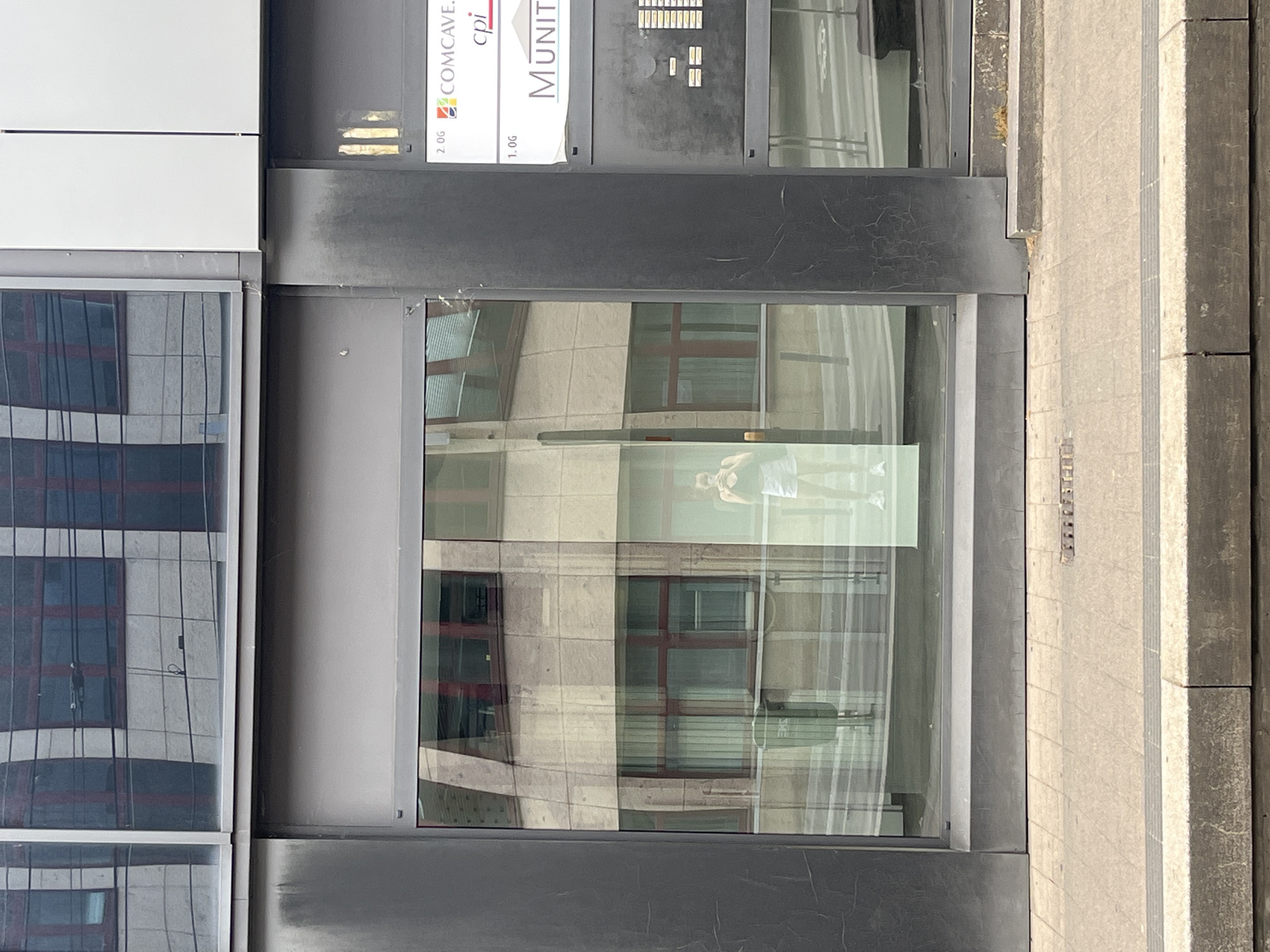 The image depicts the exterior of a building in Saarbrücken, Germany. Signs for "COMCAVE" and "MUNIT" are visible. A window reflects a nearby building. A woman is reflected in the glass window. Sidewalk tiles are on the right.