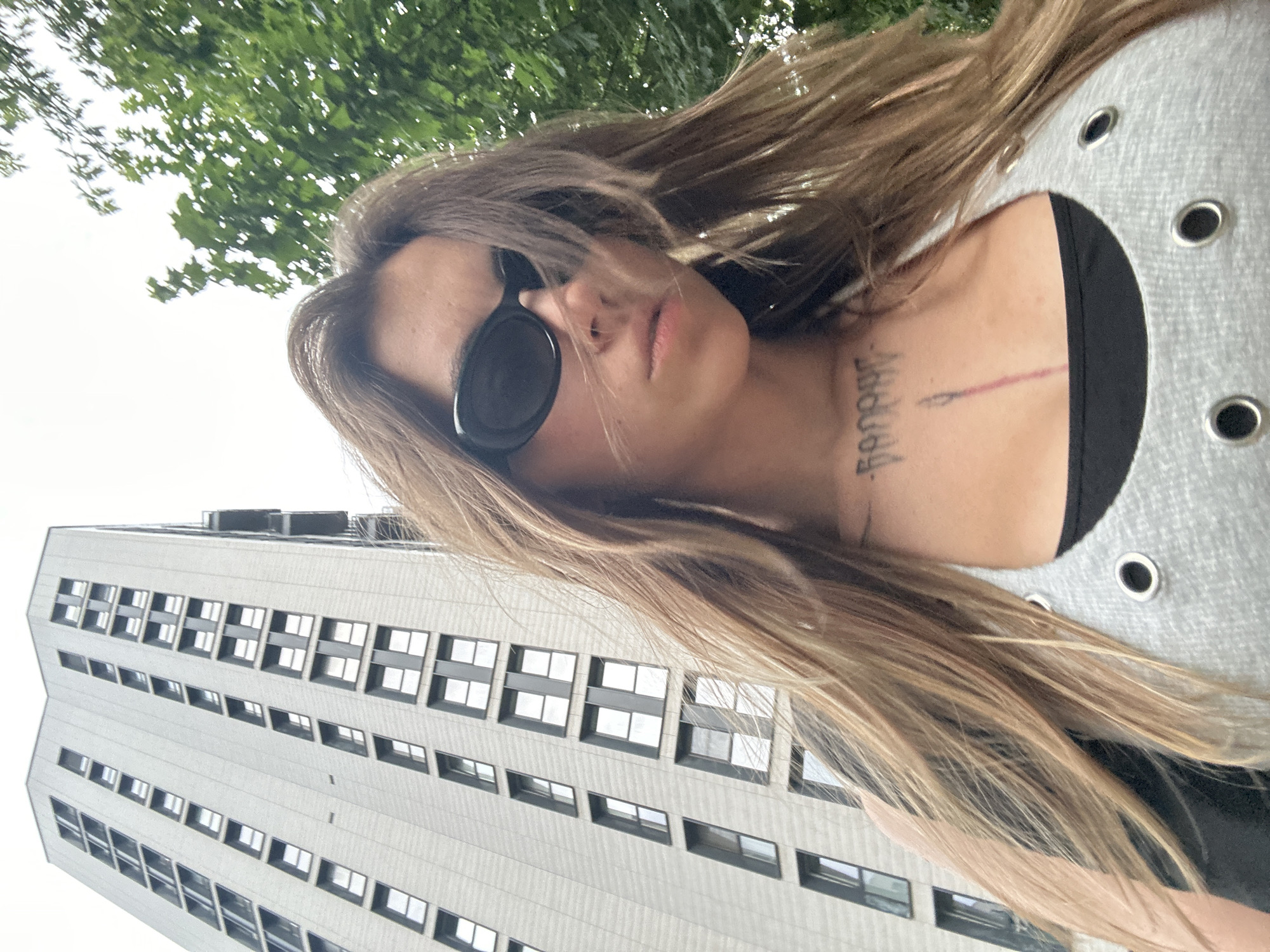 A young woman with long, blonde hair and black sunglasses is standing in front of a tall, modern apartment building. She is looking down at the camera, and her hair is blowing in the wind. The building is light gray and has many windows. There are trees in the background. The woman is wearing a gray tank top with grommets and a black shirt underneath. She has a tattoo on her chest. The image appears to be taken on a cloudy day. The location is not specified as Moscow, Russia.