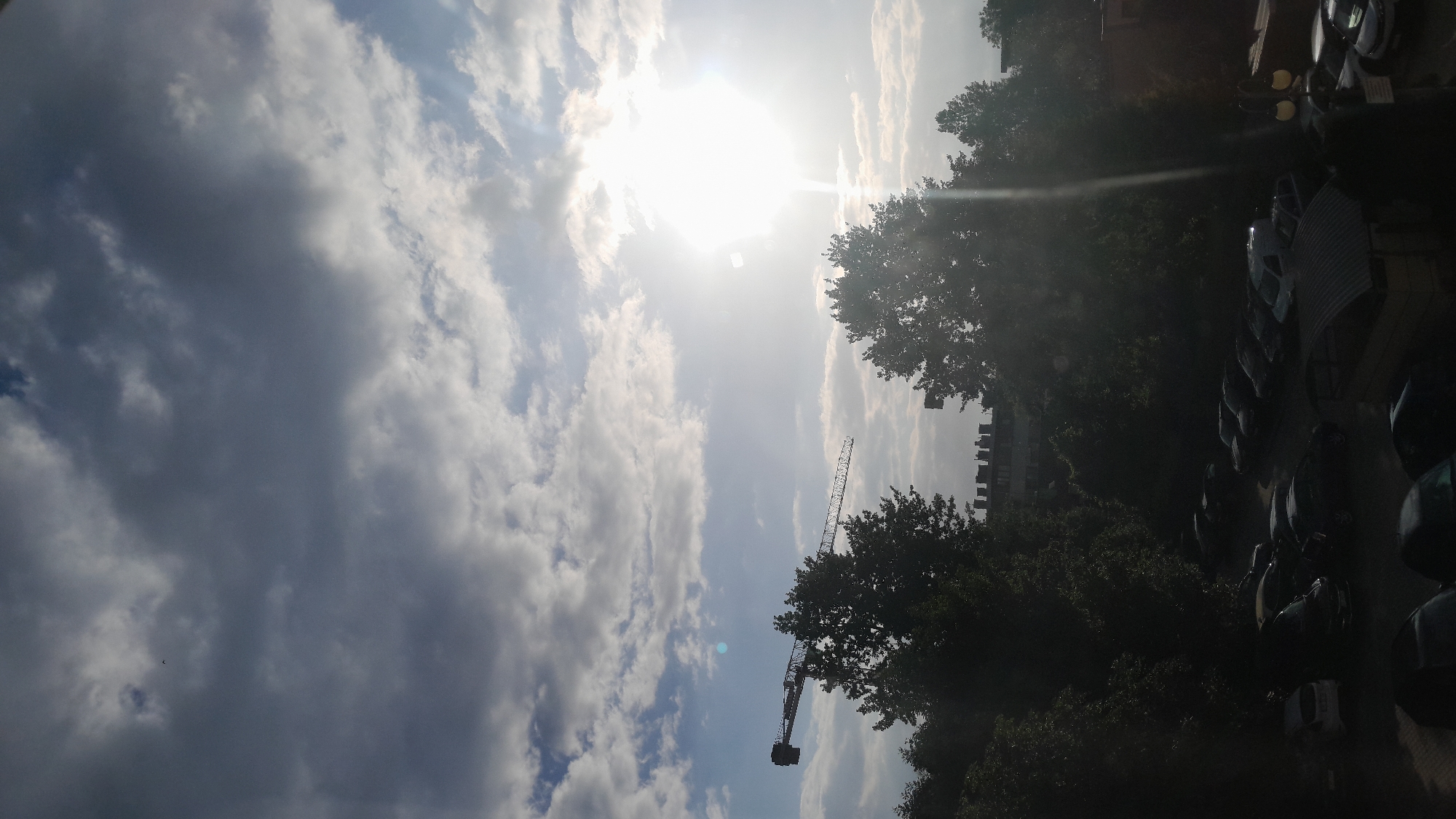 An over-the-shoulder shot captures a construction crane in the distance, silhouetted against a bright sun and cloudy sky. Trees are visible in the foreground, their dark foliage contrasting with the bright sky. Below the trees, a parking lot is filled with various cars. The scene is set in an urban environment.