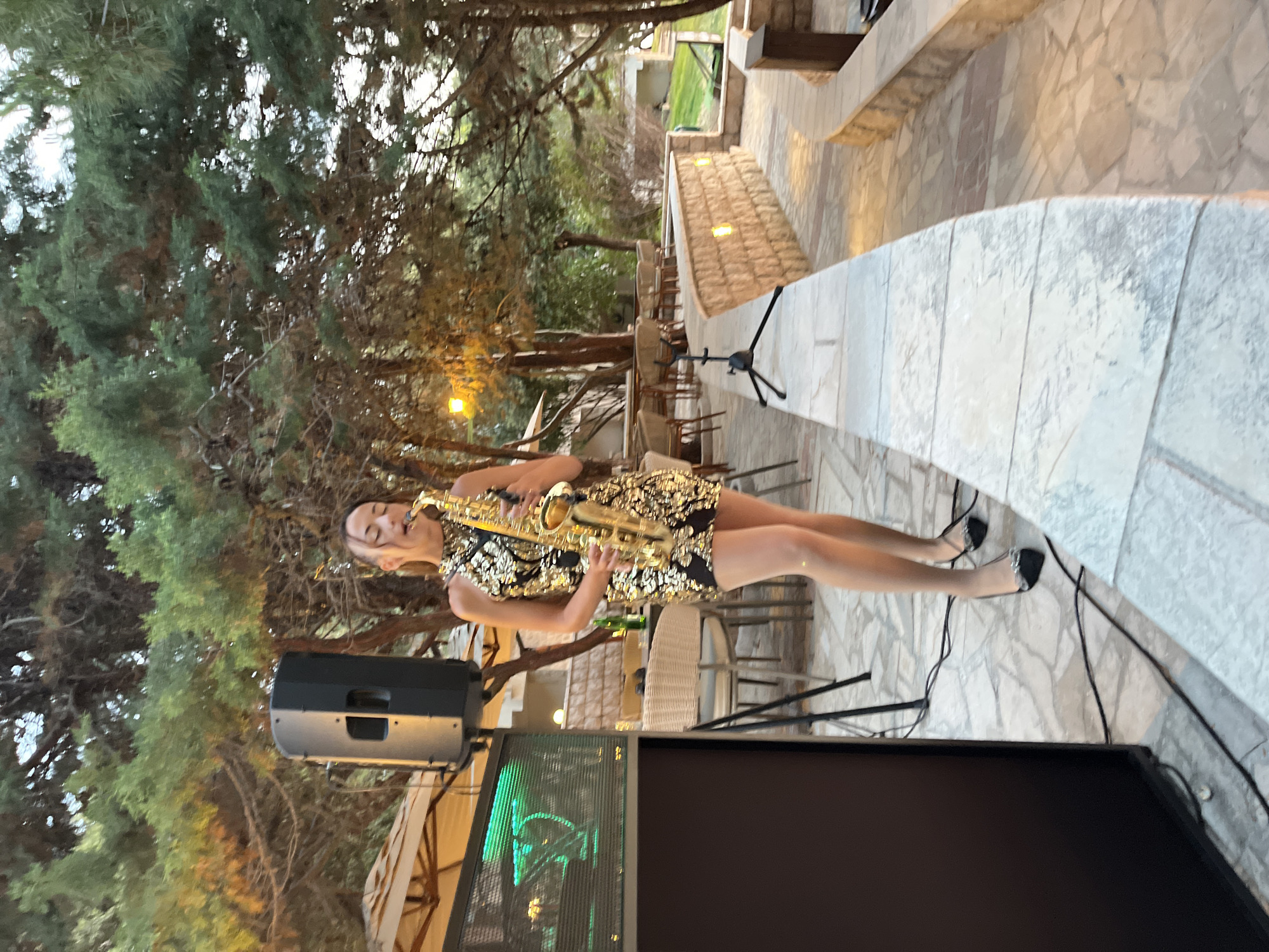 A young woman is playing a saxophone outdoors, with trees and a paved area in the background. She is wearing a black and gold sequined dress and black high heels. Her eyes are closed as she plays. In front of her is a sound system with a speaker. The scene appears to be during the day, with natural light filtering through the trees. The setting looks like an outdoor venue, possibly a restaurant or event space. The woman appears focused and immersed in her music.
