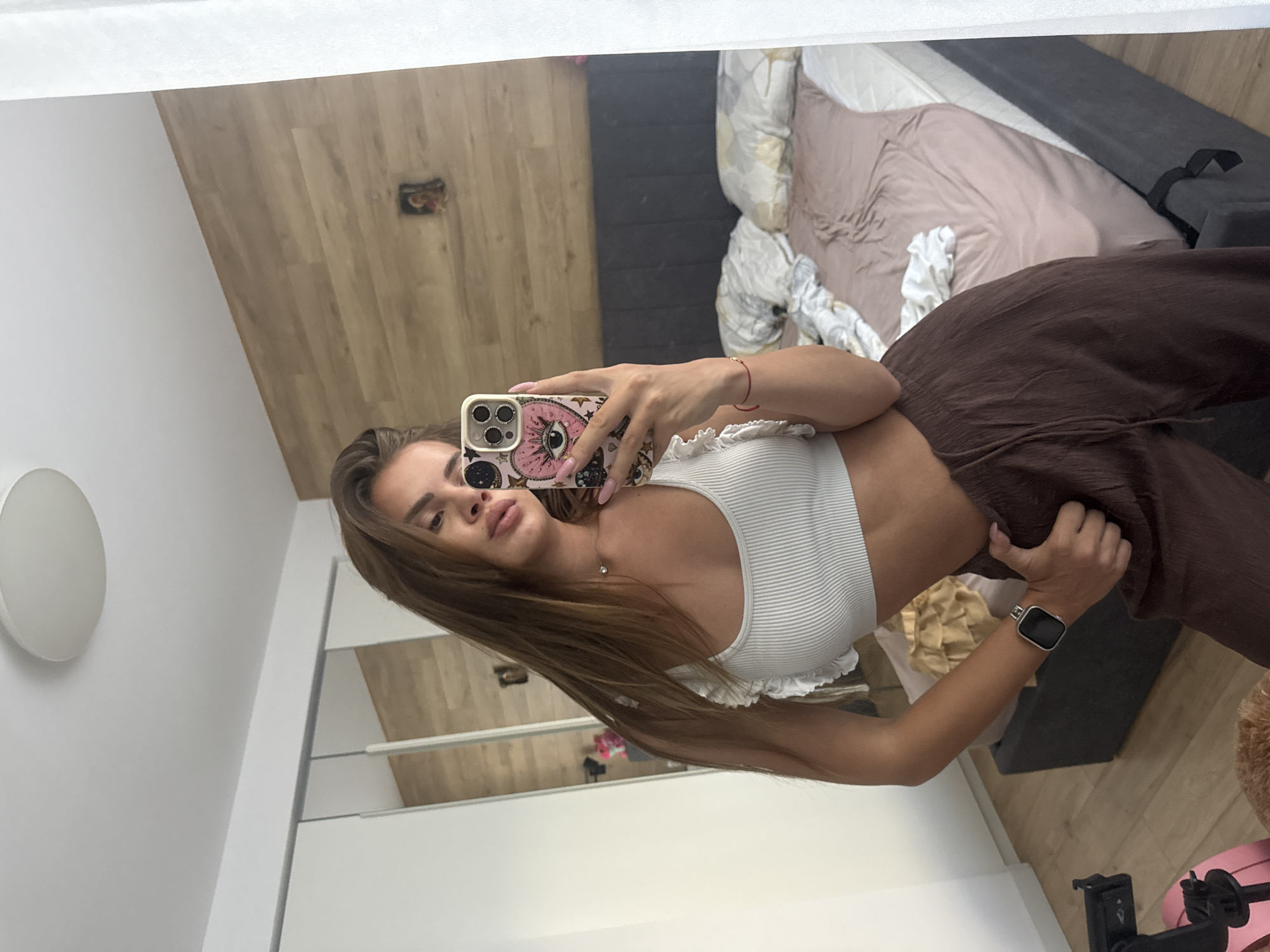 The image displays a mirror selfie of a young adult female in what appears to be a bedroom. The individual, visible from the chest to the mid-thigh, has long brown hair and is wearing a white ribbed crop top with ruffled shoulder straps and dark brown, loose-fitting pants. A square-faced smartwatch is present on her left wrist, and a red string bracelet on her right. She holds a smartphone, identifiable as an iPhone due to its triple camera array, in her right hand. The phone features a distinctive pink case adorned with an eye motif, hearts, stars, and astrological symbols. Her left hand is positioned near her waistline, seemingly in her pants.
The background reflects the interior of a modern room. The left side features a white built-in wardrobe with mirrored doors. The ceiling is white, with a round, flush-mount light fixture. On the right side, a bed with a dark grey upholstered headboard and base is visible, covered by a pinkish-brown fitted sheet and white patterned pillows. A wood-paneled wall is situated behind the bed, displaying a small framed picture or icon. The flooring appears to be light-colored wood. In the bottom-left corner, parts of a pink tripod stand and a brown furry object are discernible. No discernible visual information in the image indicates the specific city of Plovdiv, Bulgaria.
