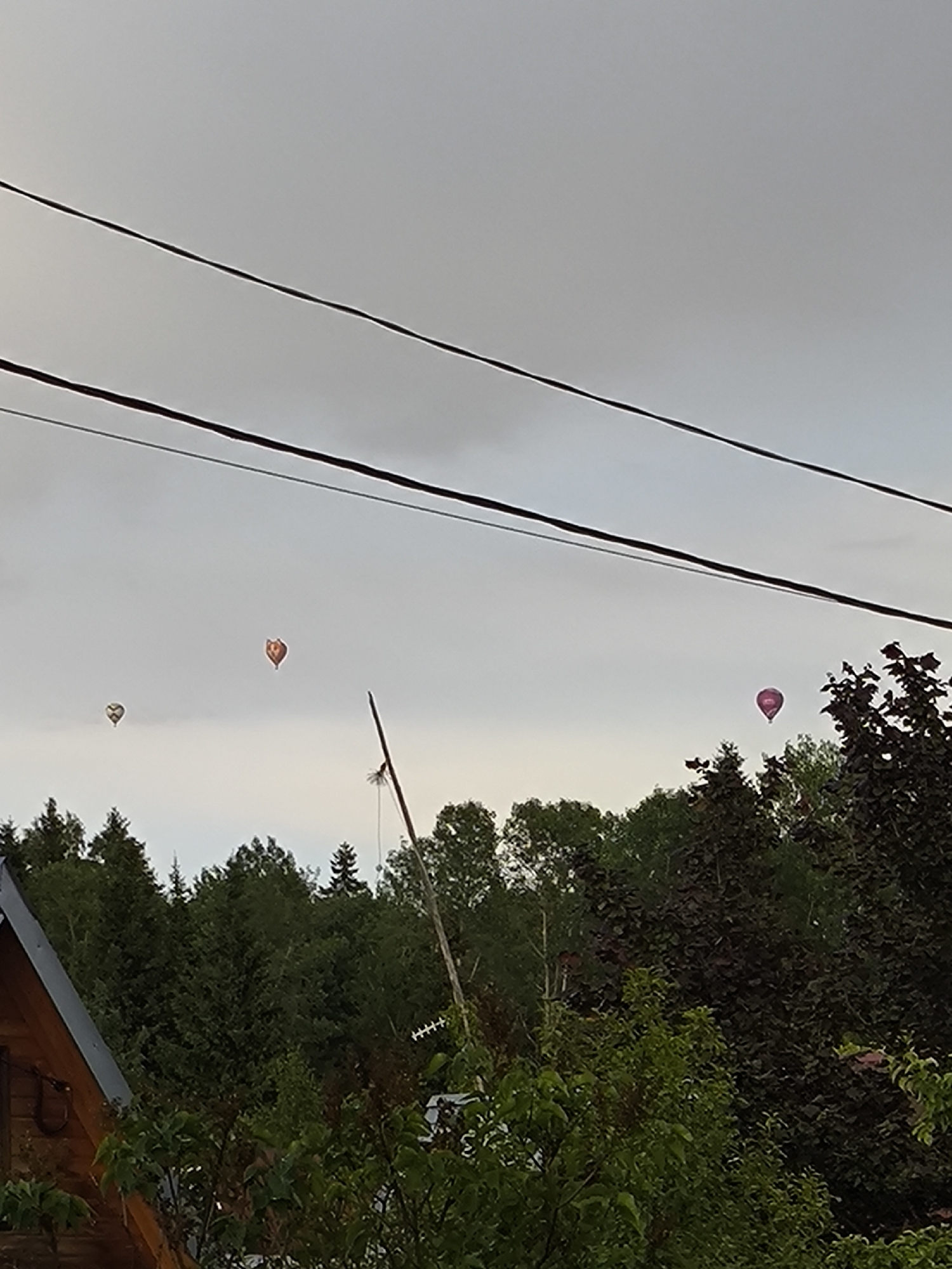 Four hot air balloons are visible in a cloudy sky above a dense forest. The balloons are of various colors, including red, orange, and purple. In the foreground, the roof of a wooden structure and some trees are visible. The scene appears to be set in a rural or suburban area.
