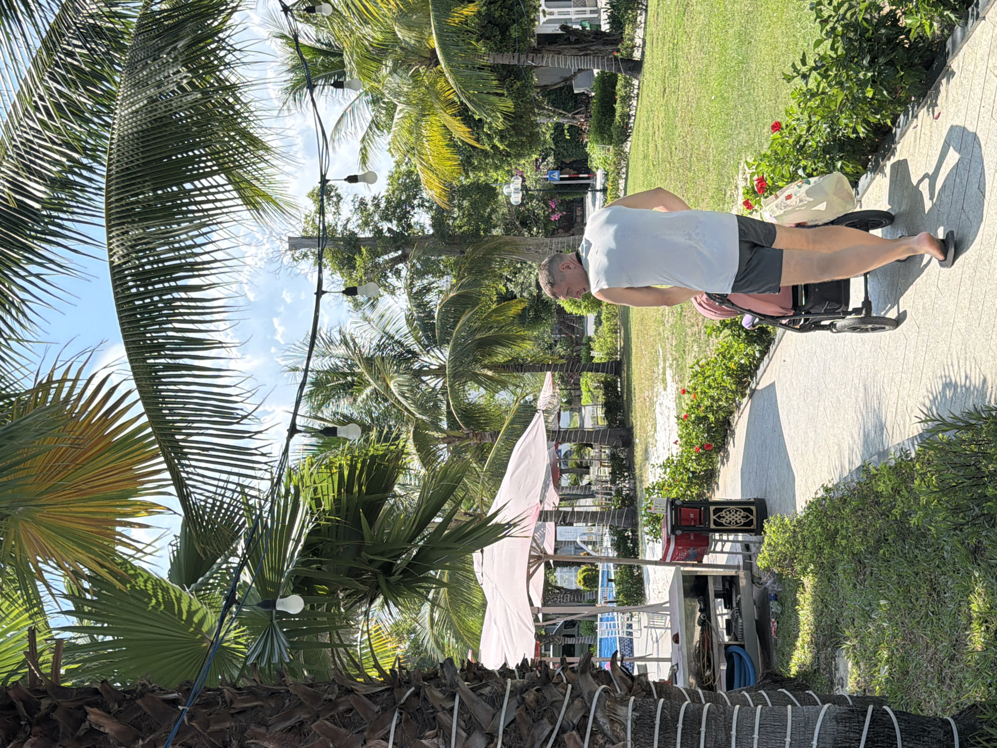 In Nam Nha Trang Ward, Vietnam, a man is pushing a stroller along a paved pathway in a sun-drenched tropical setting. He is wearing a light blue sleeveless shirt and dark shorts, with flip-flops on his feet. The stroller is a light pink color. The pathway is bordered by lush green grass and flowering bushes. Overhead, palm trees create a canopy, with string lights visible between them. In the background, there are more palm trees, some white chairs and tables, and possibly a swimming pool area with umbrellas. The sky is blue with scattered clouds.