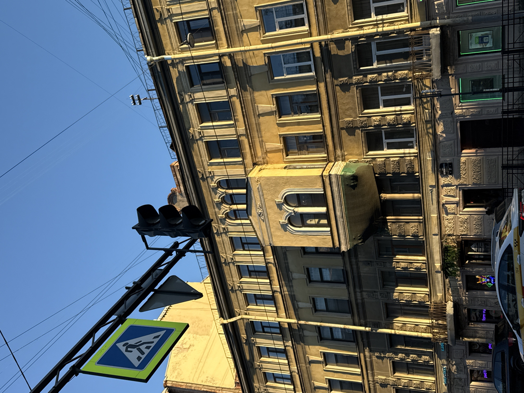 The image presents a bright, sunny street scene in Saint Petersburg, Russia, dominated by a stately, multi-story building. The building, rendered in a light yellow or beige hue, showcases classical architecture with numerous windows, ornate decorative elements, cornices, and textured panels between floors. A prominent feature is a distinctive, curved, enclosed balcony on one of the lower floors, distinguished by its arched windows.
In the foreground, a black traffic light is visible on a metal pole, beneath which hangs a standard blue and white pedestrian crossing sign with a vivid yellow-green border. Overhead, a network of electrical wires, likely for public transport like trolleybuses or trams, crisscrosses the clear blue sky.
Bright sunlight illuminates the scene, casting shadows and highlighting the architectural details. On the lower right, the front portion of a white and yellow taxi, bearing the number "764" on its side, is parked alongside a darker vehicle. The ground level of the building features several establishments with large windows, some outlined with glowing green neon. Visible text includes "tom.yu," "phc," and "pad t?" on these signs. Additionally, the partial number "1927" and indistinct Cyrillic script, possibly a street name, can be seen on the building facade near the bottom right. The scene depicts a typical busy urban environment on a clear day.