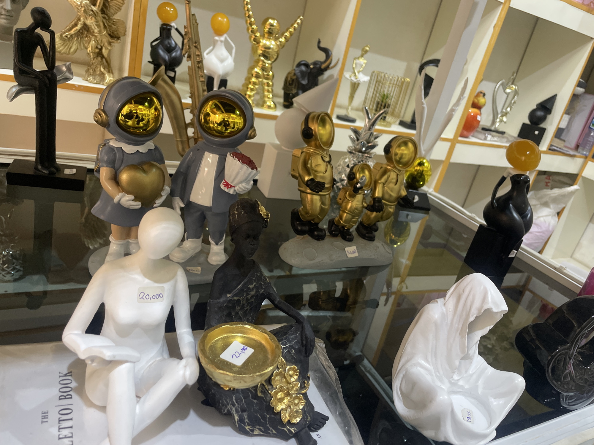 The image shows a close-up, eye-level view of a display of decorative figurines on glass shelves in a retail environment. The figurines are primarily made of ceramic or resin and include abstract human forms, astronaut figures with reflective helmets, an elephant, and golden statues. The lighting is bright, casting some reflections on the glass surfaces and the figurines. There are price tags visible on some of the items, with denominations in what appears to be local currency. The shelves are filled with a variety of other decorative items, suggesting a gift shop or home decor store. The scene appears to be indoors, with a clean and organized presentation of merchandise. The city of Jos, Nigeria, is not directly depicted in this image; the scene is a generalized retail display.