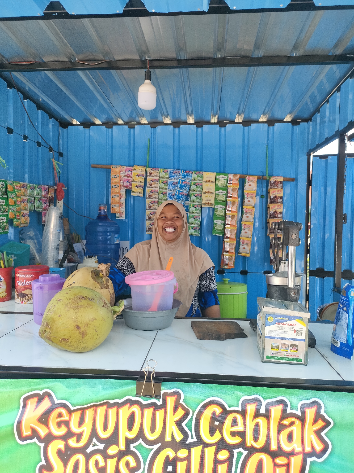 A medium shot captures a woman in a beige hijab and a blue and black patterned top smiling from behind a white counter in an outdoor food stall in Curahdringu, Indonesia. The stall has a corrugated blue metal wall and a corrugated silver metal roof with exposed wiring and a light bulb. The woman is positioned in the center of the frame.
On the counter, from left to right, are a can labeled "Khong Guan Wafer Chocolate," a purple plastic container, two whole coconuts, a pink plastic container with a pink spoon, and a gray bowl. Behind the counter, a large blue water jug is visible, along with stacks of small snack packets hanging on the wall, and a green trash bin. To the right of the woman, there is a dark gray apparatus with a handle and a silver basin, possibly for processing food, and a clear glass box labeled "KOTAK AMAL" (Charity Box). A blue and white packet is visible on the far right. A large green banner with yellow text that reads "Keyupuk Ceblak Sosis Chilli Oil" is in the foreground, partially obscured by the counter. The overall scene depicts a small, open-air food vendor.