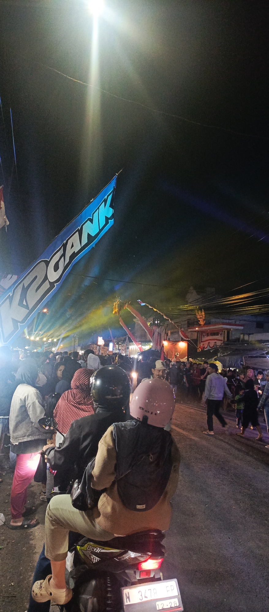 A nighttime street scene in Sekarpuro, Indonesia, is depicted, filled with a crowd of people, many of whom are on motorcycles. The atmosphere appears festive, with bright lights illuminating the area, casting beams across the dark sky. A large blue banner with white lettering, reading "K2 GANK", is prominently displayed, suggesting an event or gathering. Various stalls or temporary structures are visible along the street, indicating a possible market or festival. The focus is on the people and the lively ambiance, with individuals dressed casually in street attire. The presence of multiple motorcycles suggests transportation and mobility within the crowded space. The overall impression is one of a vibrant community event taking place after dark.
