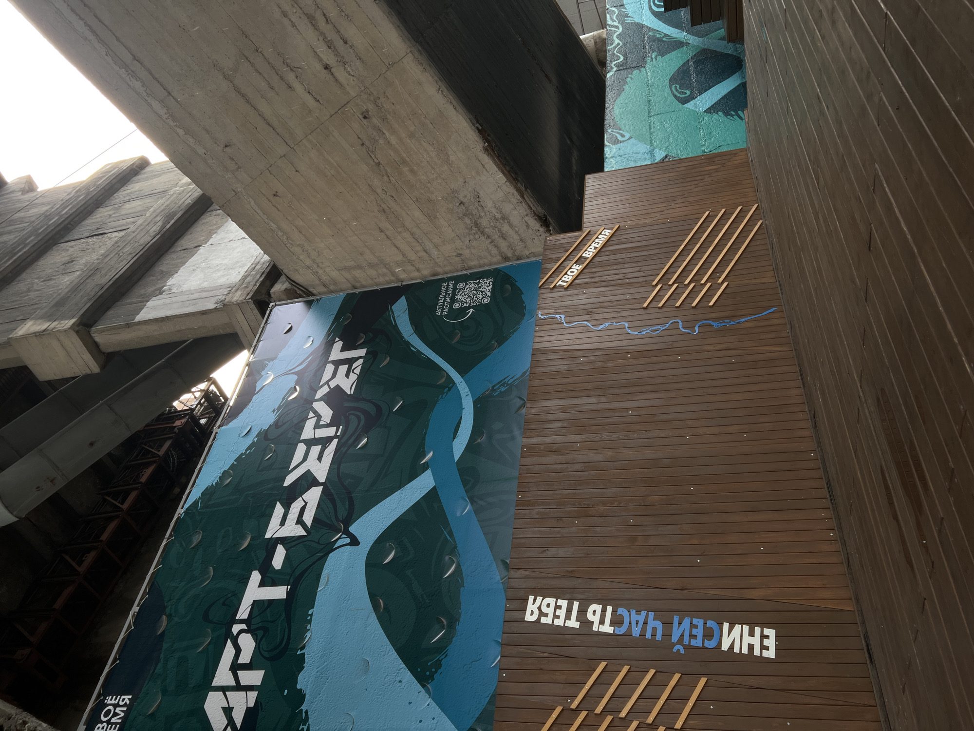 The image shows an underpass or a space beneath a bridge in Krasnoyarsk, Russia. The architecture is brutalist, with large concrete structures overhead. There is a large, dark teal and blue mural painted on a banner attached to the underside of the bridge, featuring water-like droplets and abstract waves, with white lettering that appears to say "ART-EXPO" or similar. To the right of the mural, a QR code is visible next to text that reads "АКТУАЛЬНОЕ РАСПИСАНИЕ" (current schedule) and "ТВОЕ ВРЕМЯ" (your time).
Below the banner, there is a wall clad in vertical dark brown wooden planks. White and blue lettering on this wall reads "ЕНИСЕЙ ЧАСТЬ ТЕБЯ" (Yenisei, part of you) vertically on the left, and "ТВОЕ ВРЕМЯ" (your time) diagonally on the right. There are decorative wooden slats arranged diagonally on the wooden wall, and a thin blue wavy line painted to resemble a river.
The ground in the foreground is a wooden deck, also laid with planks, which appears wet with puddles. To the right, there is a continuation of the space with more graffiti on a concrete wall, featuring green and blue abstract designs. The overall impression is of an urban public art installation or event space in an unconventional location.