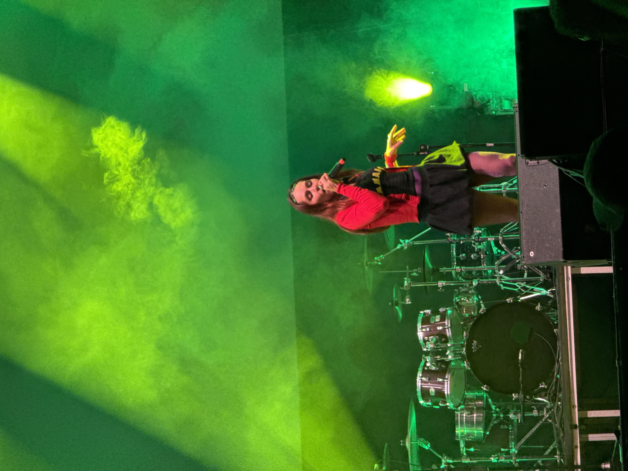A female singer is performing on stage at a concert, possibly in Vilanova i la Geltrú, Spain. She is holding a microphone and gesturing with her right hand. She is wearing a red long-sleeved shirt and a black skirt. The stage is lit with green lights and smoke effects. A drum kit is visible behind the singer, and a large speaker is on the right side of the frame.