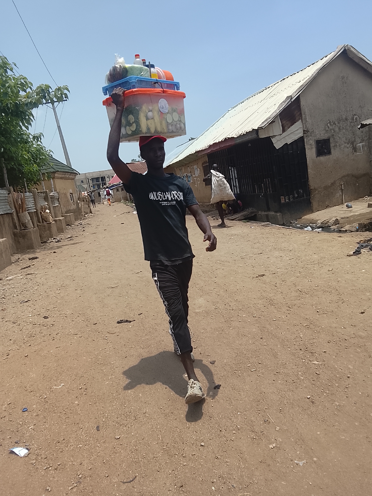 A man walks down a dusty road in Abuja, Nigeria, balancing two plastic containers on his head. The top container is blue, while the bottom is orange. Both appear to hold food or beverages. The man is wearing a black t-shirt, black pants with a white stripe down the side, worn sneakers, and a red baseball cap. The road is unpaved and lined with buildings. Some buildings have corrugated metal roofs. A few other people are visible in the distance. The sky is clear and bright.