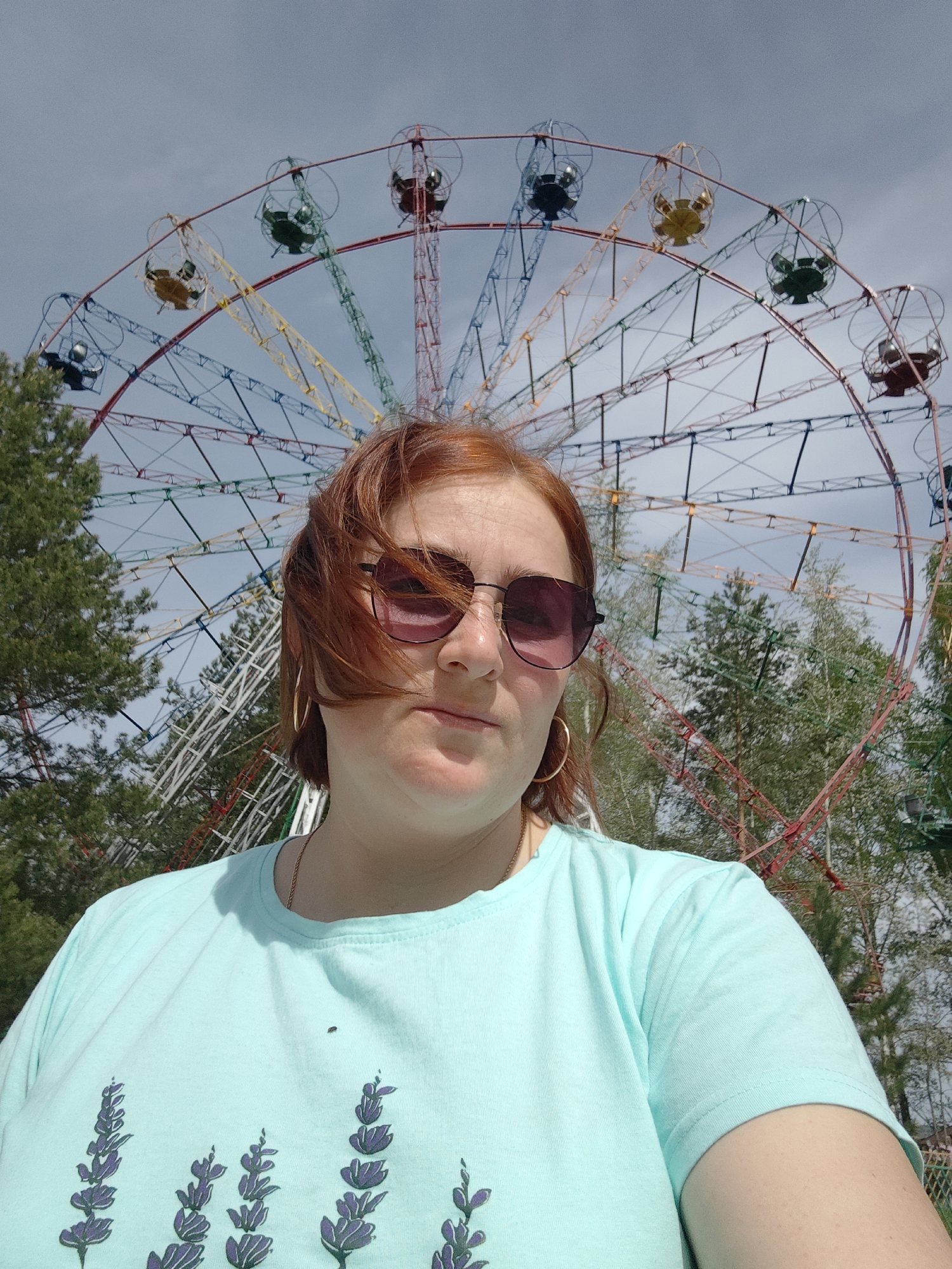 A woman stands in front of a Ferris wheel in Chaikovsky, Russia. The Ferris wheel has cars in various colors, including red, yellow, green, and blue. Trees are visible in the background, adding to the outdoor setting. The woman wears sunglasses and a light blue t-shirt with a purple floral design. She has red hair and is wearing gold hoop earrings. The sky is partly cloudy.