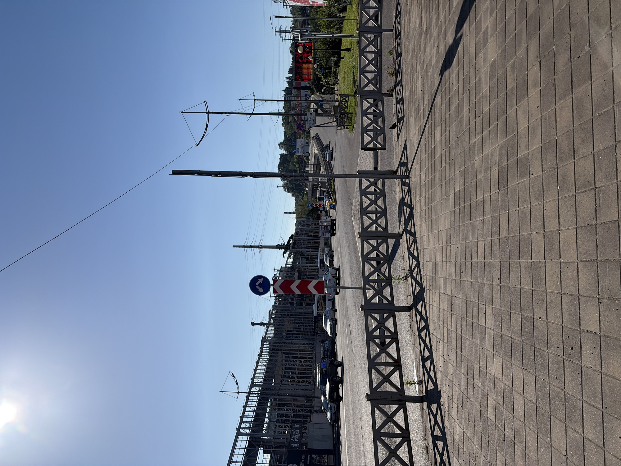 The image depicts an urban or semi-urban street scene on a bright, sunny day, likely in Sochi, Russia. The perspective is oriented along a road, with a wide, tiled sidewalk on the right and a street with parked cars and construction on the left.
In the foreground and to the right, a sidewalk made of rectangular gray paving stones stretches into the distance. A sturdy metal barrier, featuring a repeating X-pattern, runs along the edge of the sidewalk, separating it from the road. This fence casts long, distinct shadows across the pavement, indicating either early morning or late afternoon sun.
The midground features a multi-lane road where several vehicles, including dark cars and a white truck/van, are either parked or moving. On the left side of the road, extensive scaffolding and partially built structures suggest a large construction project or an industrial site. Beyond this, a green hillside covered in trees and some distant buildings are visible.
The clear, bright blue sky dominates the upper portion of the image. Numerous utility poles and power lines crisscross the sky. The glare of the sun is visible in the upper left quadrant.
Notable details include a blue circular traffic sign with two white arrows, likely indicating a mandatory direction or a roundabout, and a red and white chevron sign typically used to mark a sharp curve or obstacle. A large red billboard is partially visible in the background, along with a smaller sign below it that includes Cyrillic text, most notably "СОЧИ" and numbers such as "90, 110," confirming the location. Another sign near the construction site reads "PORADDA" and "STREET FOOD." No people are explicitly visible, suggesting a focus on the urban infrastructure.