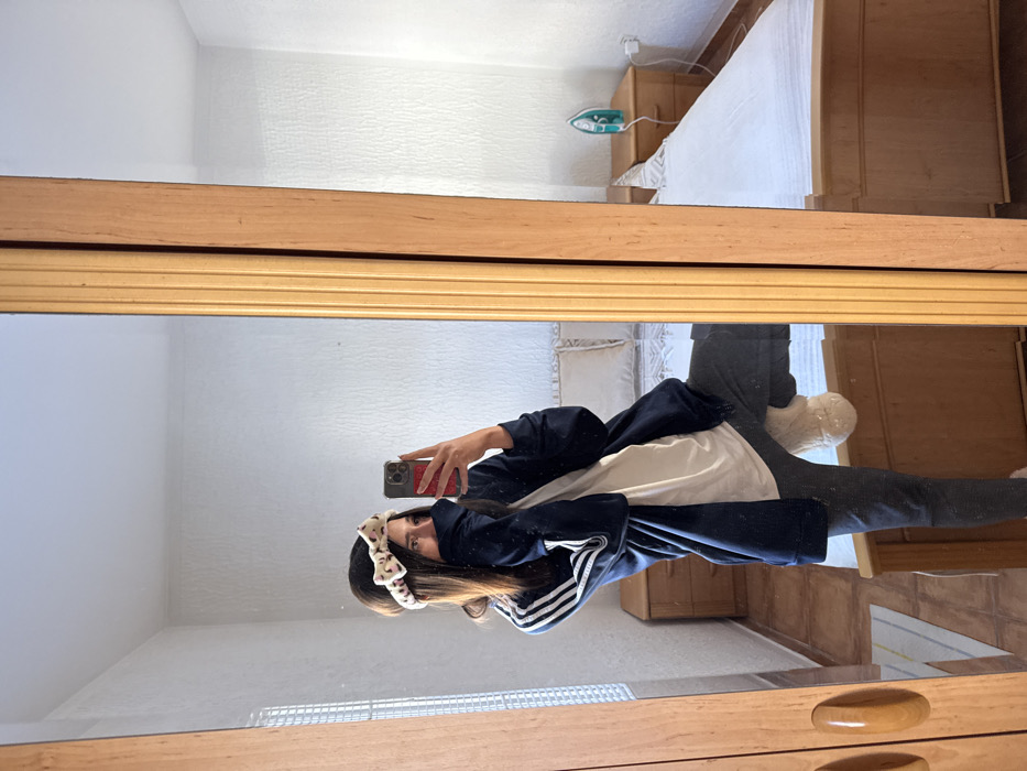 The image depicts an indoor scene within a residential room in Zankovići, Montenegro. A single individual is captured in a mirror self-portrait.
The individual, with brown hair and a cream-colored headband featuring dark patterns, is wearing a navy blue Adidas track jacket with white stripes on the sleeves, a white t-shirt, dark trousers, and cream-colored fluffy slippers. The right hand holds a dark-colored smartphone with a red case, angled to capture the reflection. The individual's face is mostly obscured by the jacket's collar, with only the eyes and forehead visible. The left leg is bent at the knee, with the foot raised.
The mirror has a wooden frame, featuring a lighter, ribbed section on the right and a smoother, darker section on the left.
The reflected background reveals a bedroom setting. A bed with a light wooden frame, white mattress, and white bedding is visible, flanked by two light wooden nightstands. On the right nightstand, a turquoise iron rests on a white ironing board. A white electrical outlet with a plugged-in white cable is present on the wall above this nightstand. The walls are white and textured. A window with white Venetian blinds is visible on the left side of the reflection. The floor is light-colored wood or laminate, with a small striped rug (white, grey, yellow) partially visible on the floor to the left of the reflected individual's feet. The room is brightly lit, consistent with natural light.