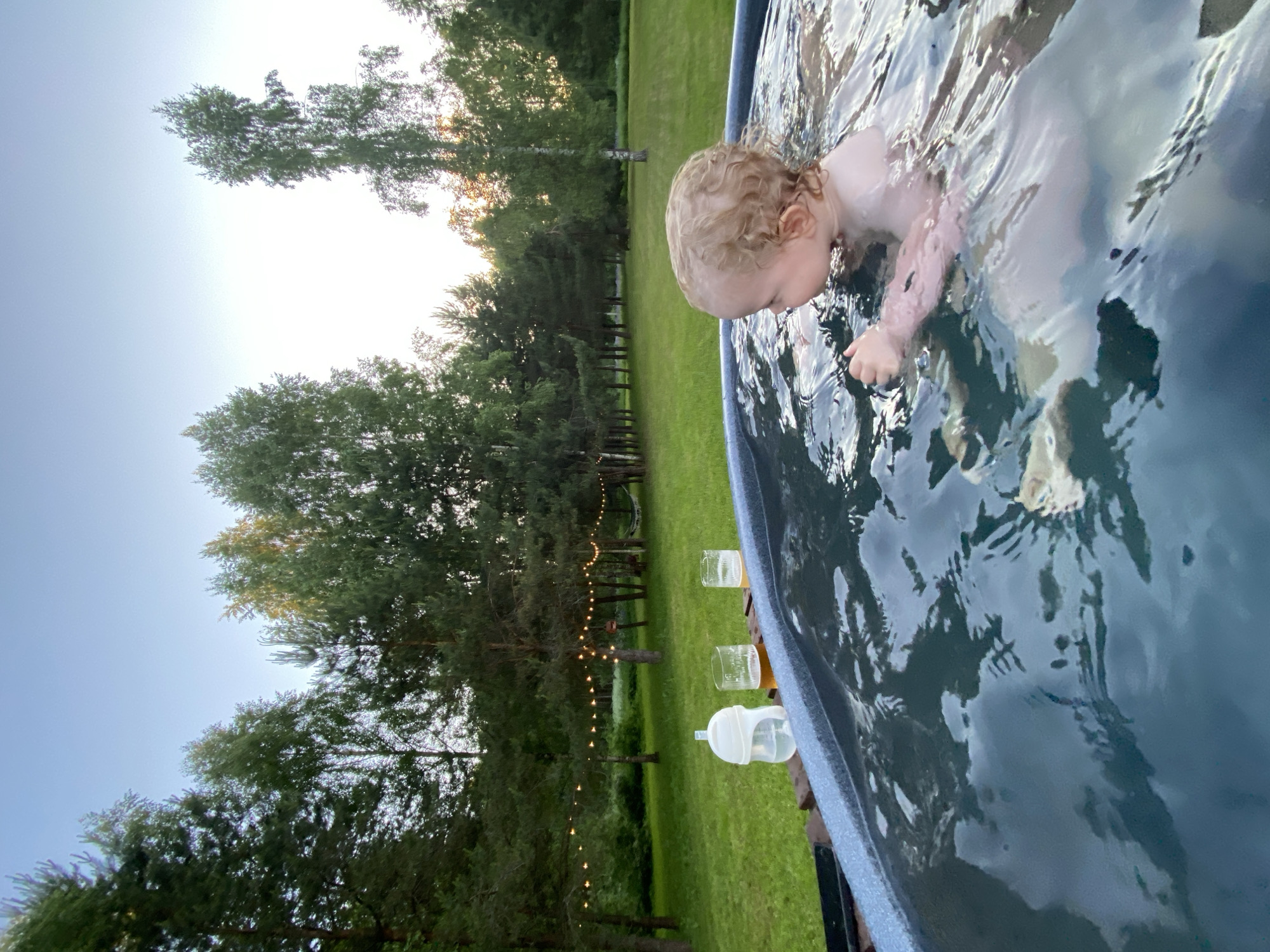 An outdoor scene in Копнино, Russia, depicts a child in a dark blue hot tub during twilight hours. The child, with light curly hair, is positioned in the upper right quadrant of the hot tub, facing right and partially submerged with their head, shoulders, and arms visible above the water. On the edge of the hot tub, positioned on a wooden surface, are three beverages: a white baby bottle containing clear liquid, and two clear glasses filled with an amber-colored liquid, one with visible foam.
The background features an expansive, well-maintained green lawn extending to a dense line of trees, including both coniferous and deciduous species. A string of warm-toned fairy lights is draped between several trees in the mid-ground, illuminating the area. The sky above the tree line transitions from light blue at the zenith to a brighter, yellowish-white hue near the horizon, characteristic of sunset or sunrise. Reflections of the sky and surrounding trees are visible on the surface of the water within the hot tub.