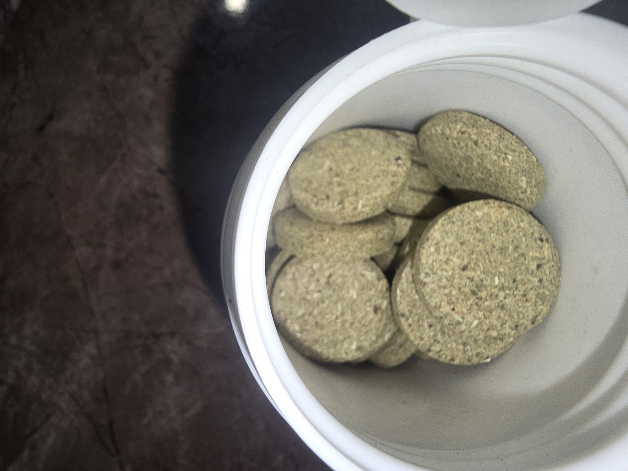The image is a close-up, top-down view of a white plastic container filled with numerous small, flat, circular, greenish-brown compressed tablets. The tablets appear to be made of organic material with visible flecks and textures. The container is partially open, revealing the stacked contents. The background is dark and out of focus, showing a textured surface. The image does not depict any identifiable urban environment or location within Ulan-Ude, Russia.