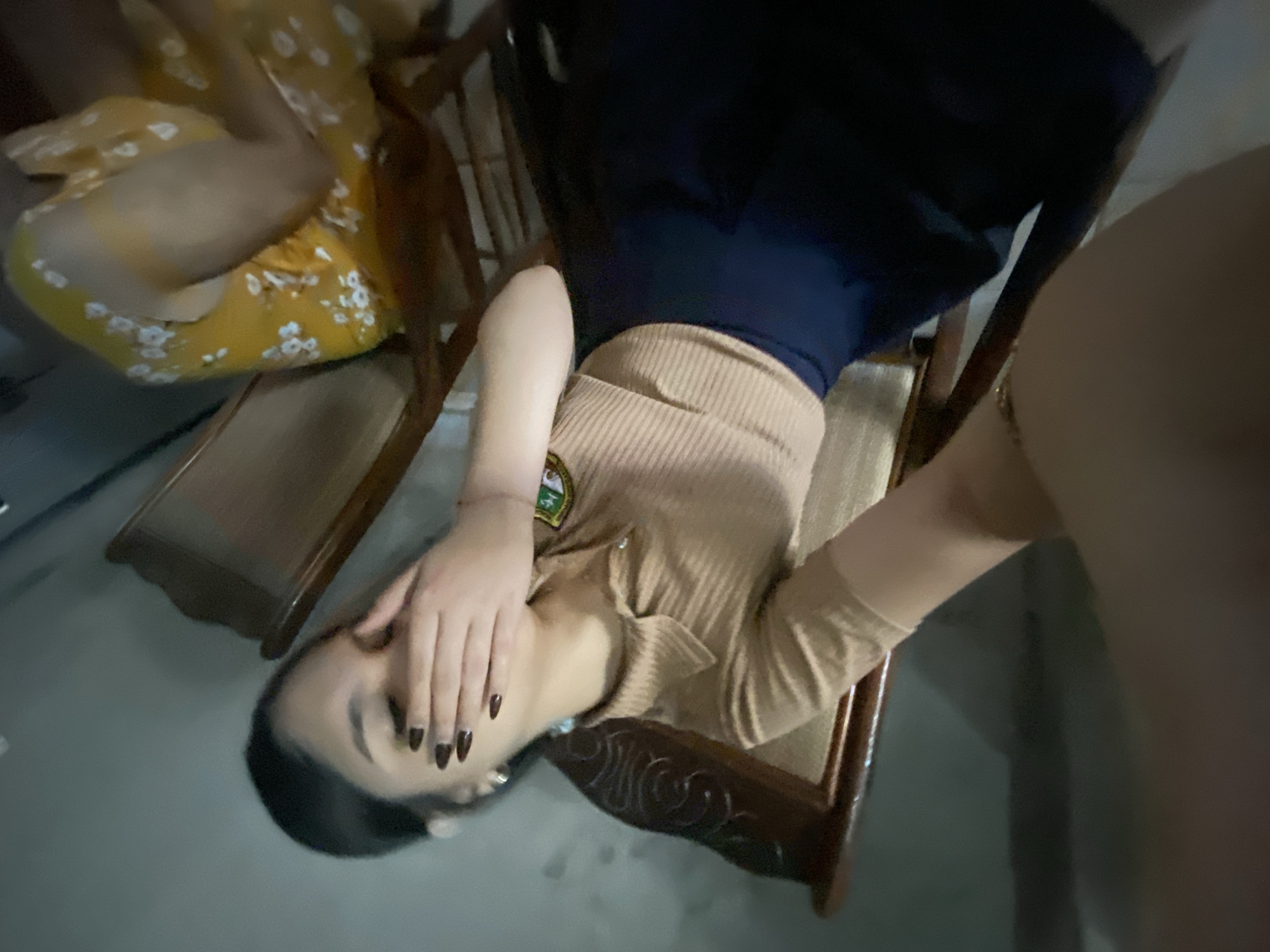 A person is sitting on a wooden chair, covering their face with their hand. They are wearing a beige top and a dark blue garment. Another person in a yellow floral dress is partially visible in an adjacent chair. The setting appears to be indoors, with a view of a gray floor and a wall. The location of Cotoperíz, Venezuela, is not discernible from the image.
