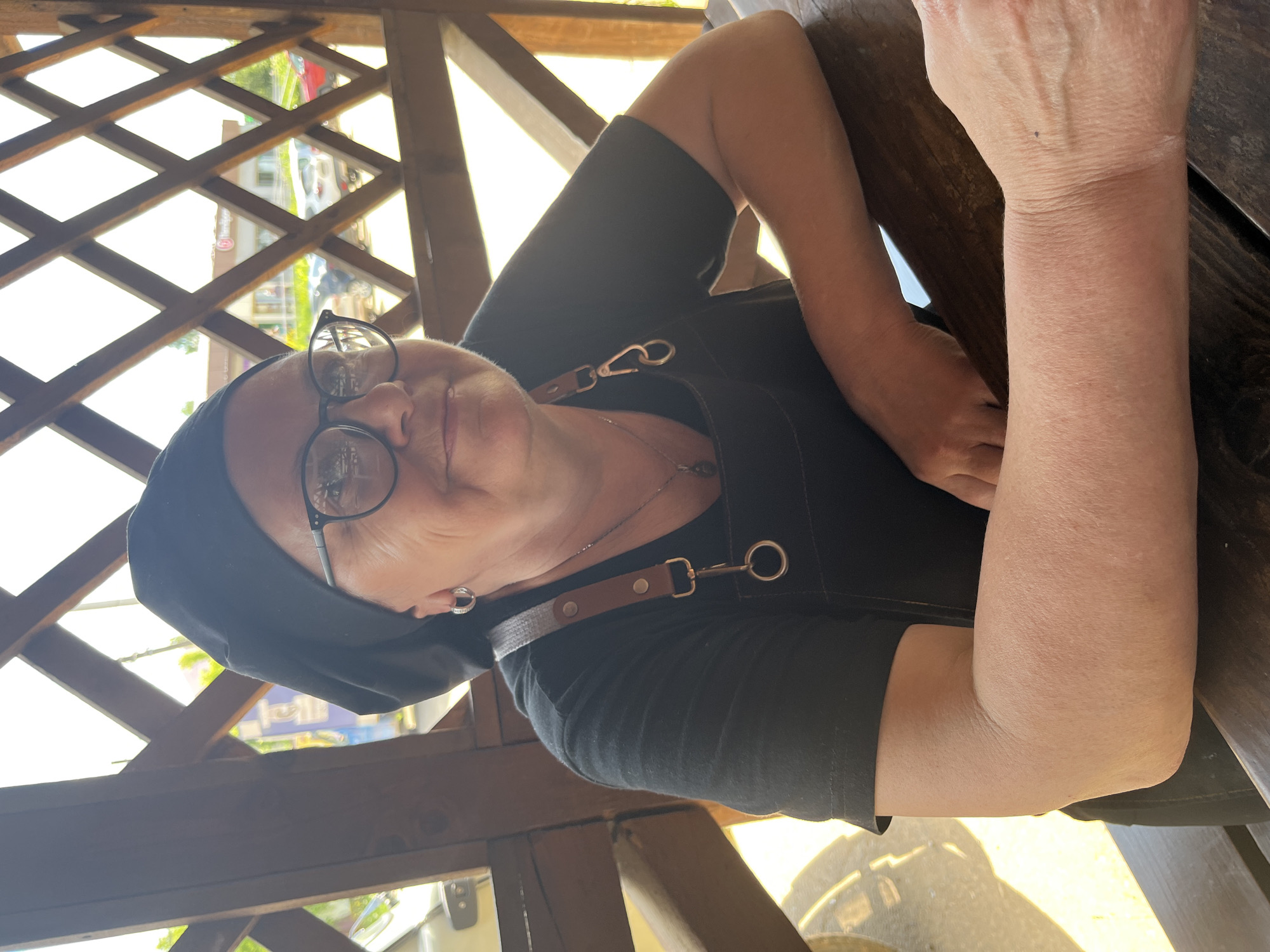 A medium shot shows a woman wearing a black chef's hat, black t-shirt, and black apron. She is seated at a wooden table, leaning forward with her arms resting on it. The woman has glasses and appears to be smiling slightly. The background consists of a wooden lattice structure, with glimpses of trees and buildings through the openings. The lighting suggests it is daytime, possibly outdoors. The location is identified as Fokino, Russia.