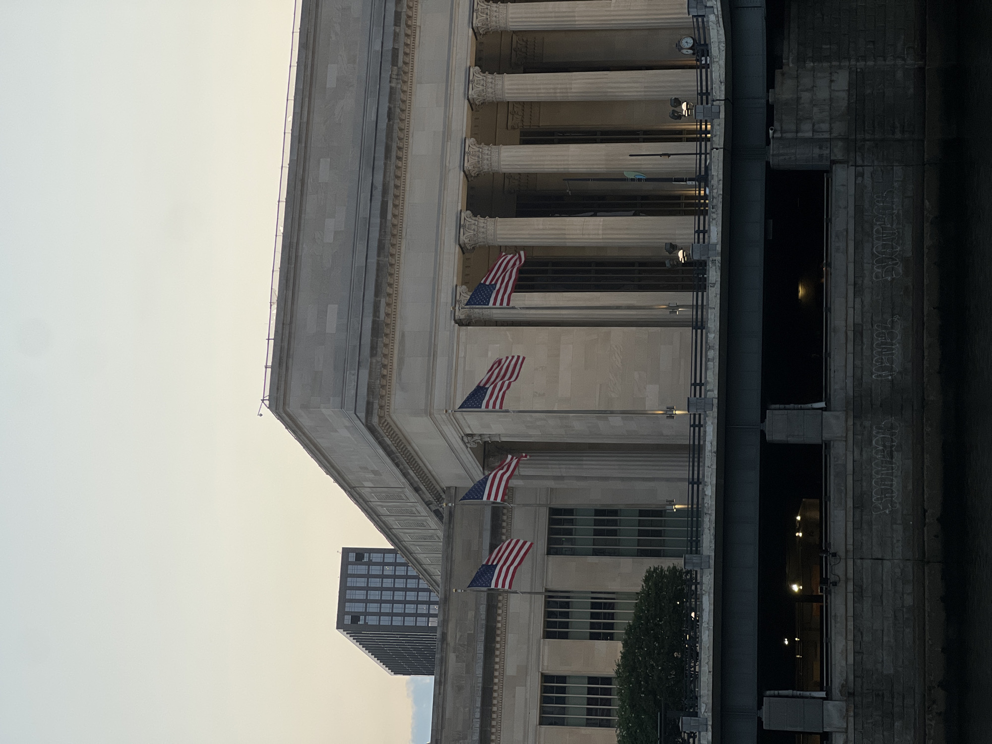 The image shows the facade of 30th Street Station in Philadelphia, United States of America, a grand Beaux-Arts building constructed of limestone. The building features a prominent colonnade with Corinthian columns and a classical cornice. Three American flags are displayed on flagpoles in front of the station. A modern, dark-colored skyscraper with numerous windows is visible in the background, to the left of the station. A bridge passes in front of the lower portion of the station, obscuring the street level. The underside of the bridge is illuminated by artificial lights, and graffiti is visible on the stone wall supporting the bridge. The sky above is a pale, overcast hue, suggesting either early morning or late evening. There are no people visible in the image.