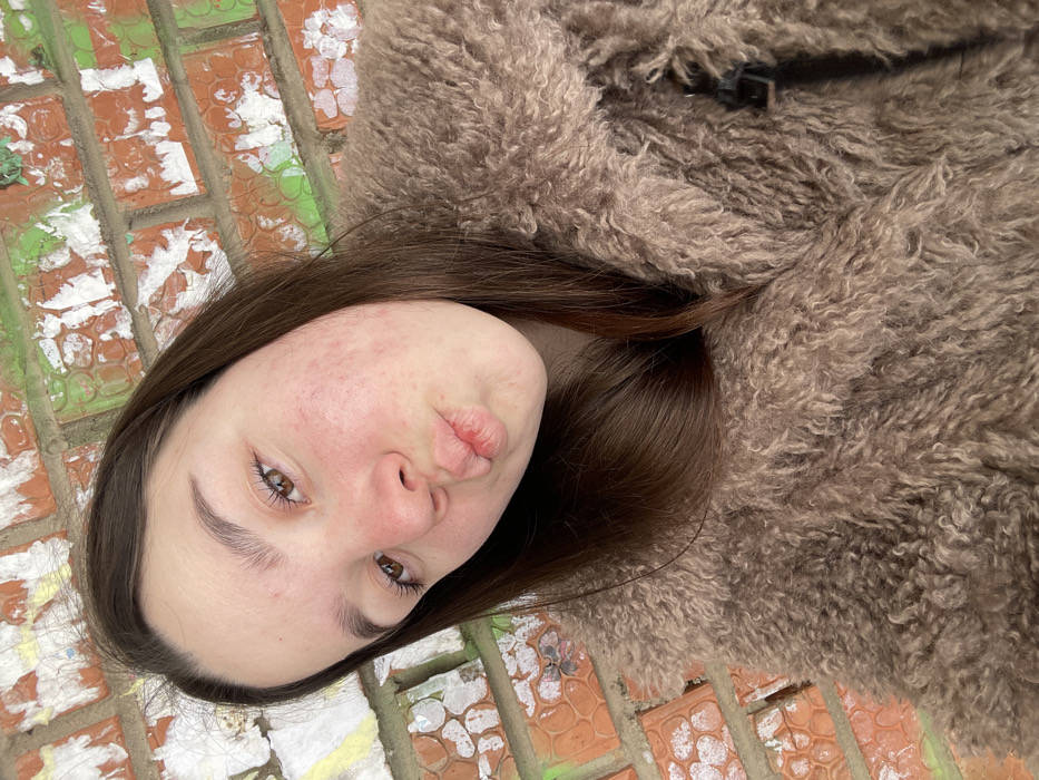 A young woman lies on a brick surface, covered in paint. She's wearing a fuzzy, brown jacket. She has light brown hair and brown eyes. Her face shows some acne and redness. She's making a puckered lip expression, appearing playful. The angle suggests a selfie, capturing her from slightly above.