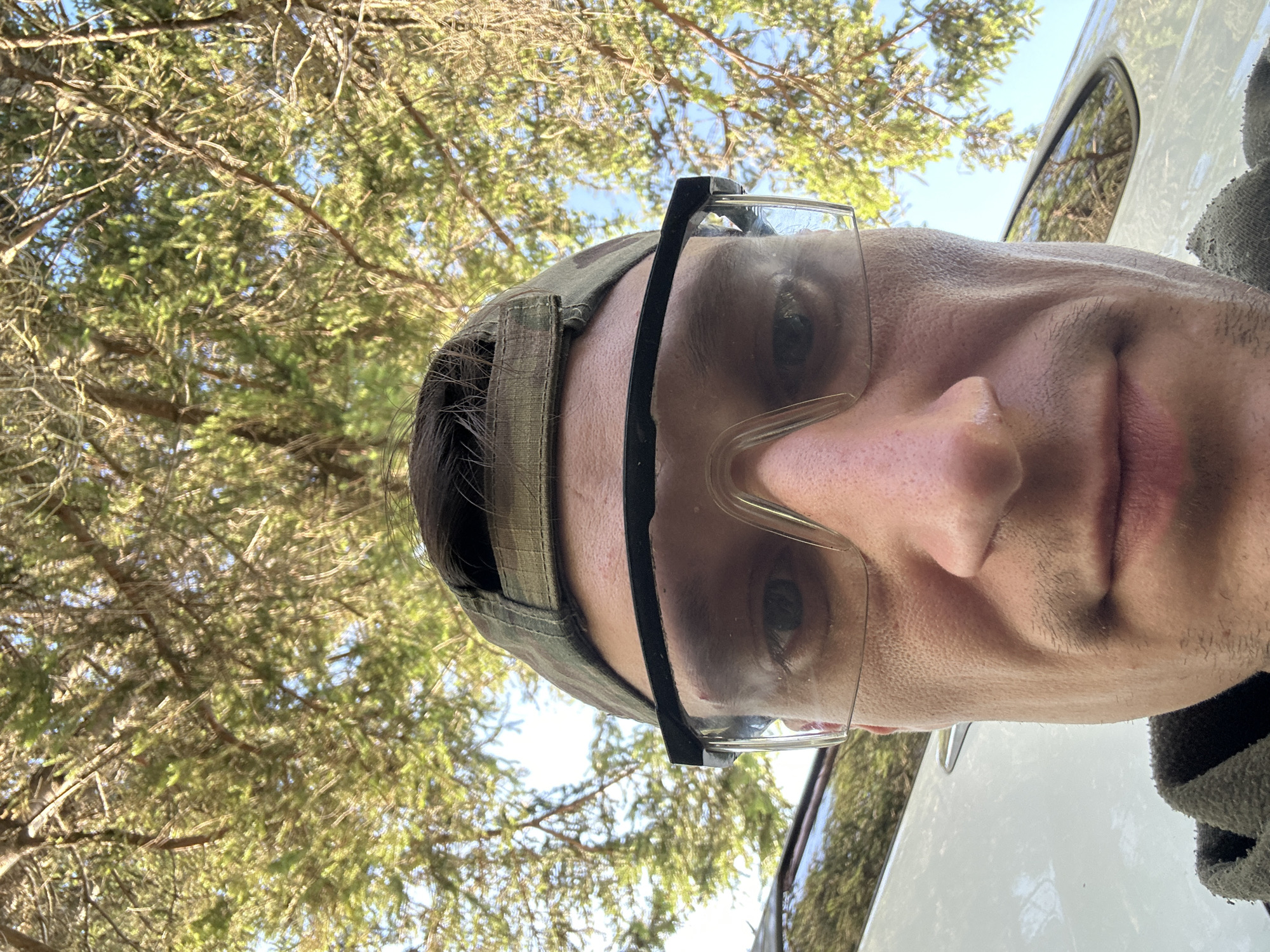 This is a close-up, low-angle, outdoor photograph featuring a person's face and upper body. The subject is wearing a camouflage baseball cap and clear safety glasses. The person appears to be looking directly at the camera. The background consists of green foliage from trees and a bright blue sky, suggesting it is daytime with clear weather. To the right of the person, a portion of a white vehicle is visible, including a car door and window. The camera is positioned below eye level, looking upwards at the subject. The overall scene suggests the person is outdoors, possibly in a natural setting or engaged in an outdoor activity. No visible text is present in the image.