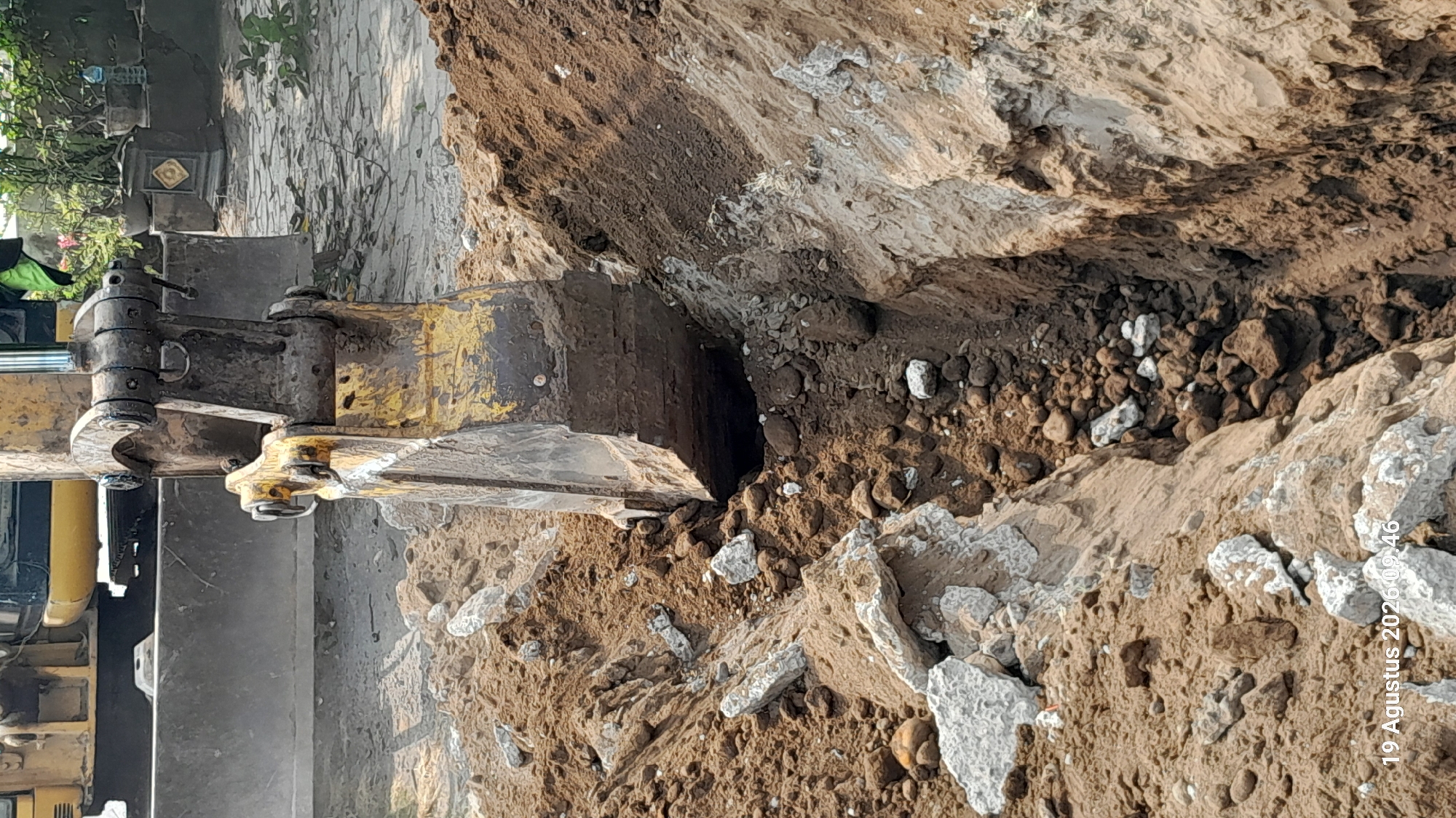 In Takeran, Indonesia, a close-up shot captures an excavator digging a trench. The excavator's yellow bucket, stained with mud and dirt, is partially submerged in the freshly dug earth. Large chunks of concrete and smaller rocks are scattered around the excavation site, indicating demolition or construction work. The trench itself is narrow and deep, with loose soil and debris lining its sides. The background is blurred but suggests an outdoor environment with some greenery and structures. A timestamp in the lower left corner reads "19 Agustus 2026 09:46".