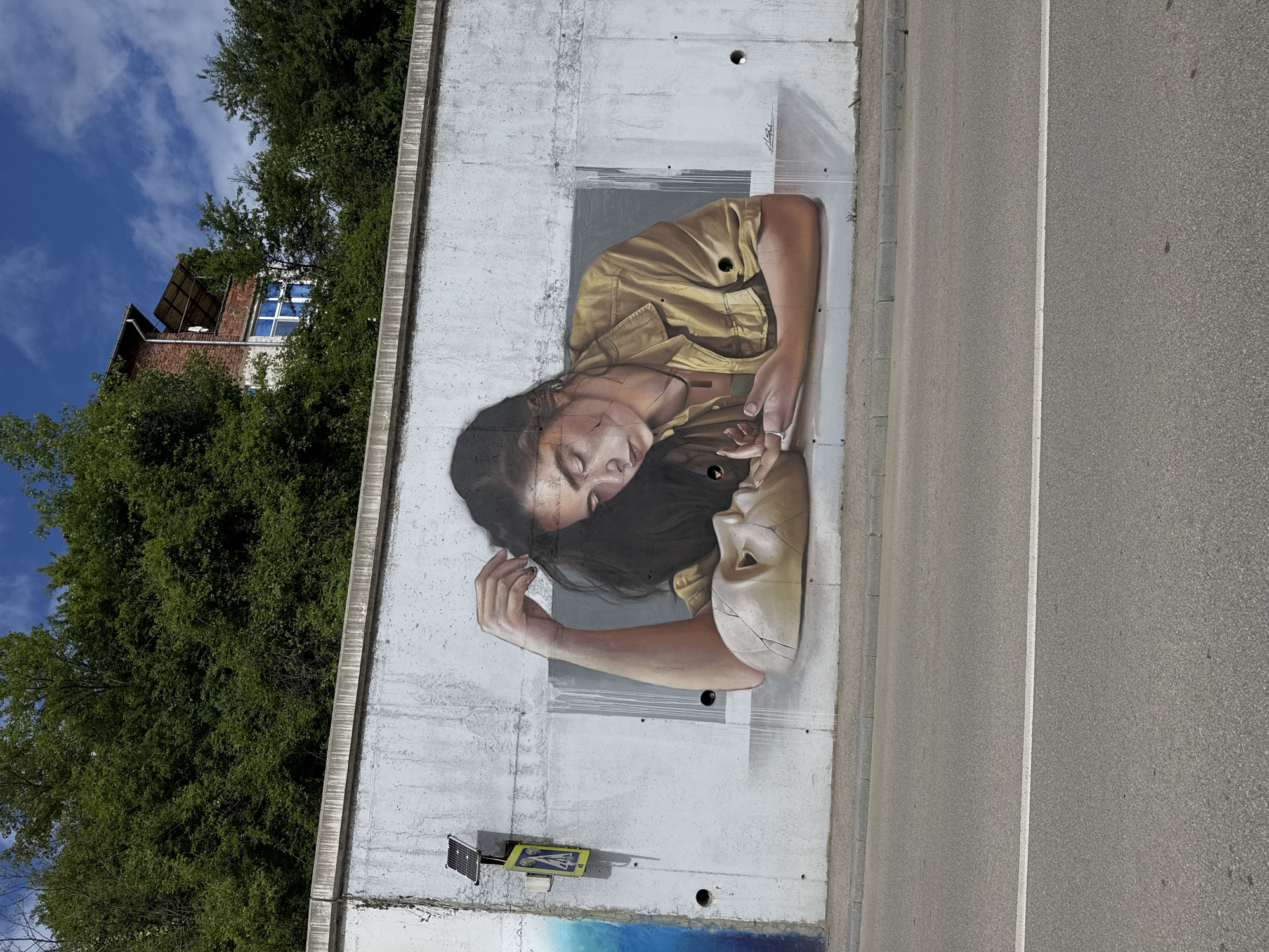 The image depicts a mural on a concrete wall in Gabrovo, Bulgaria. The mural portrays a person with dark hair, wearing a khaki-colored shirt, resting their head near a cracked mask. The wall is adjacent to a road with a white line marking the lane. Behind the wall, there are trees and a building visible under a blue sky with some clouds. A small solar panel and a piece of blue-painted art are located near the bottom of the wall. The overall scene suggests an urban setting with public art.