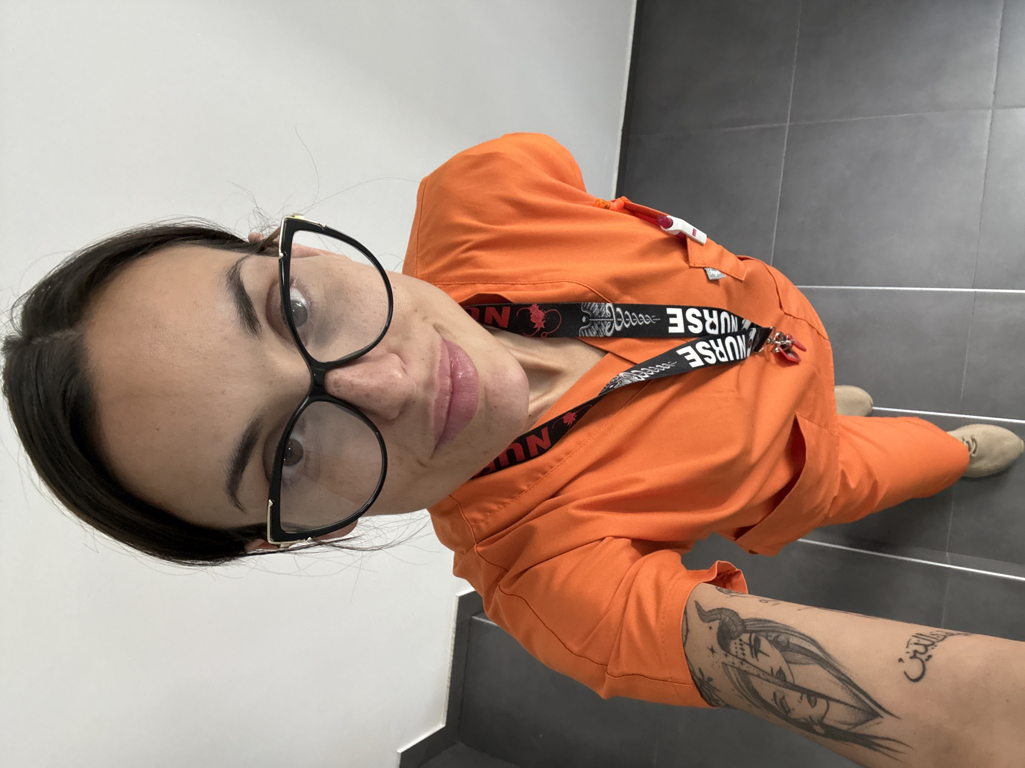 A young adult woman with dark hair and wearing eyeglasses takes a selfie. She is dressed in orange scrubs with a lanyard around her neck that reads "NURSE." She has tattoos on her left forearm. She stands on a staircase with gray tile flooring. The background includes white walls. There is no information available that would place this scene within the city of Pleven, Bulgaria.