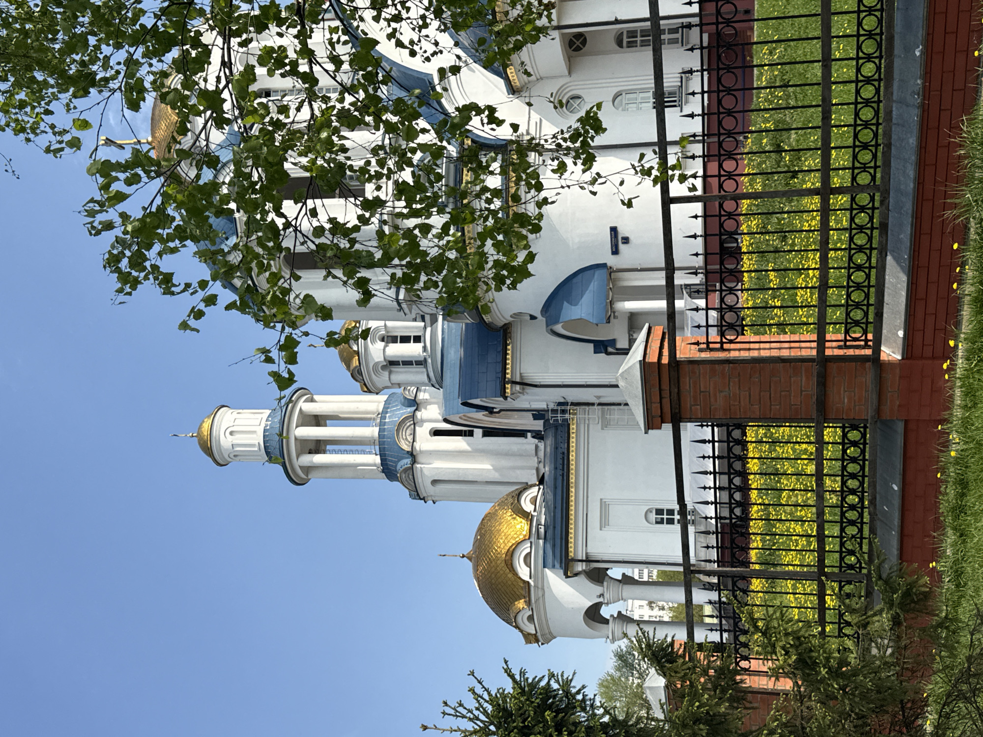 The image shows a church with white walls and blue and gold domes. The church is partially obscured by trees. A black iron fence with brick pillars runs along the side of the church, separating it from a grassy area with yellow flowers. The sky is clear blue. Based on the architecture, this is likely an Orthodox church in Moscow, Russia.