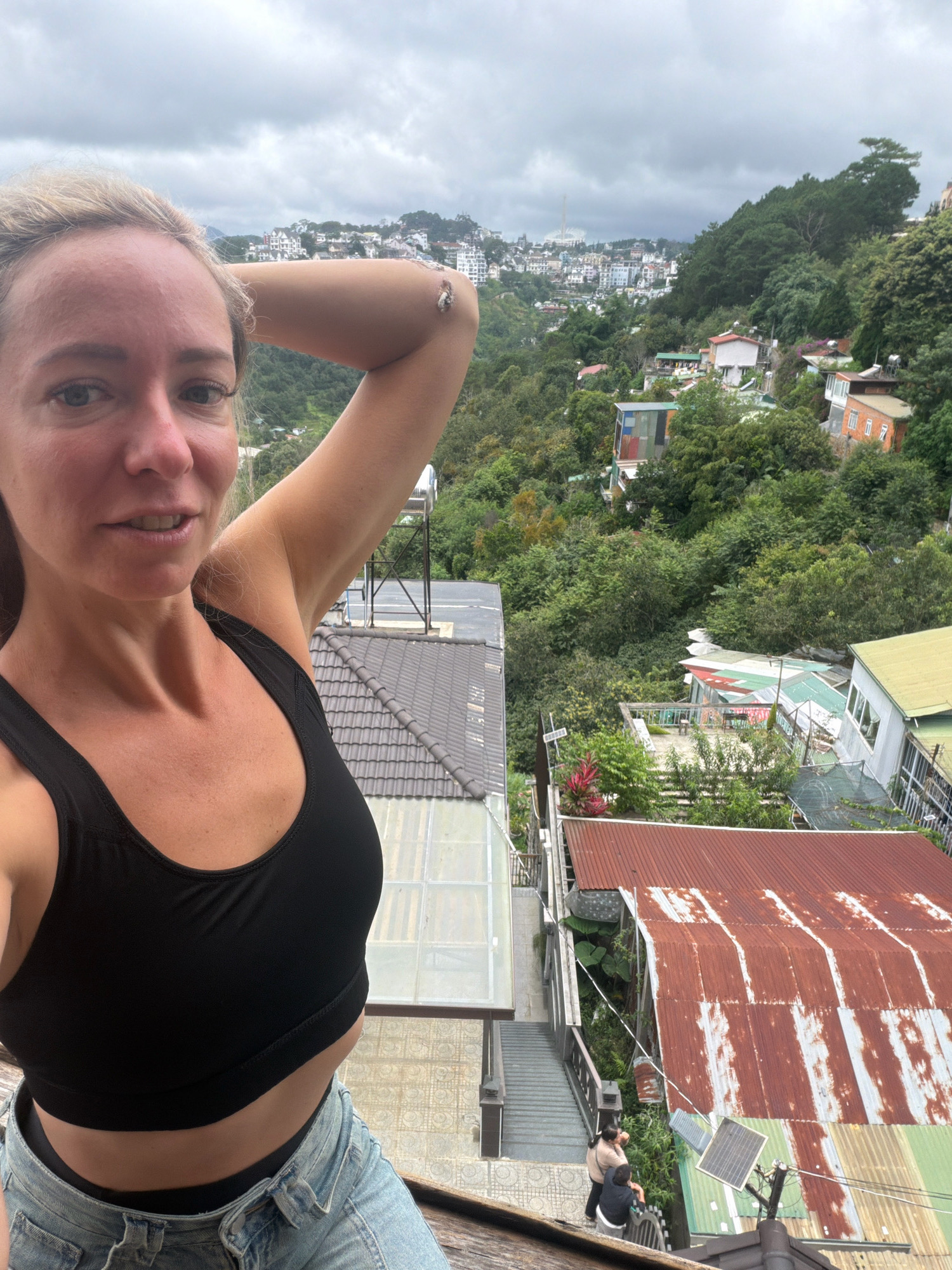 A blonde woman in a black sports bra and denim shorts takes a selfie outdoors. She is positioned in the foreground, with her left arm raised and bent behind her head, revealing a view of Da Lat, Vietnam, in the background. The city is built on a hilly terrain, with numerous houses and buildings clustered together amongst lush green trees. The sky is overcast with a dull gray hue. In the mid-ground, rooftops of various houses are visible, some made of corrugated metal, others of tiled material. There are also some solar panels and water tanks. A staircase with a railing is visible on the right side, leading down to a pathway where two individuals are standing. The overall scene depicts an urban landscape characterized by its hilly topography and dense vegetation.