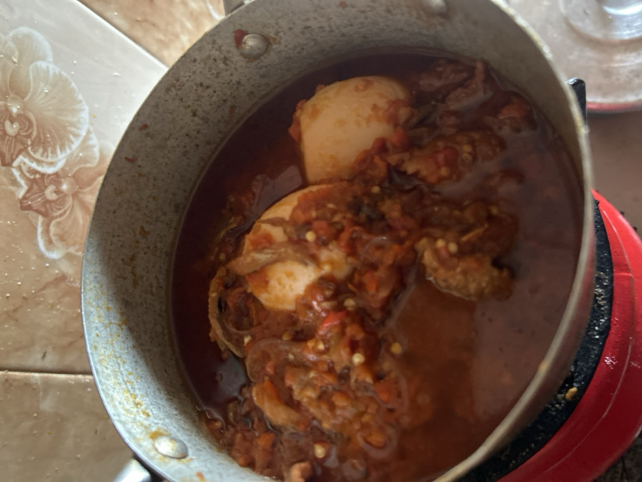 A close-up, top-down image depicts a light-grey metallic cooking pot containing a reddish-orange stew, positioned within an indoor kitchen setting in Egbe, Nigeria. The stew includes at least two whole, peeled, light-colored eggs, irregular brown pieces consistent with meat or fish, and visible red particulate matter throughout the sauce. The pot, exhibiting wear, rests on a partially visible red and black circular heating element. The background consists of light-brown tiled walls, one tile displaying a white, raised floral relief pattern in the upper right quadrant. No individuals are present.